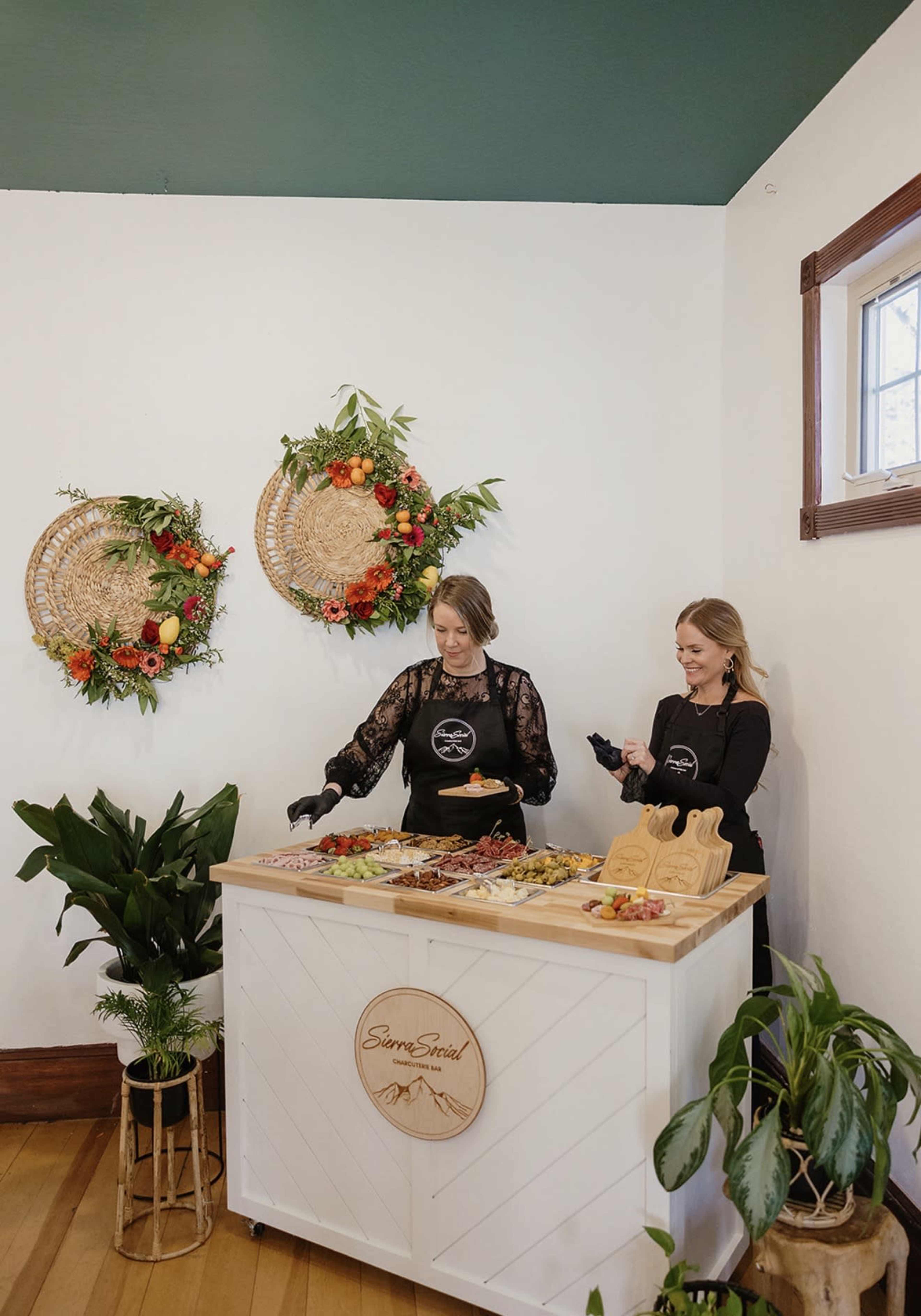Two women in black aprons stand behind a wooden food display table adorned with various appetizers, while decorative wreaths made of flowers and fruits hang on the wall.
