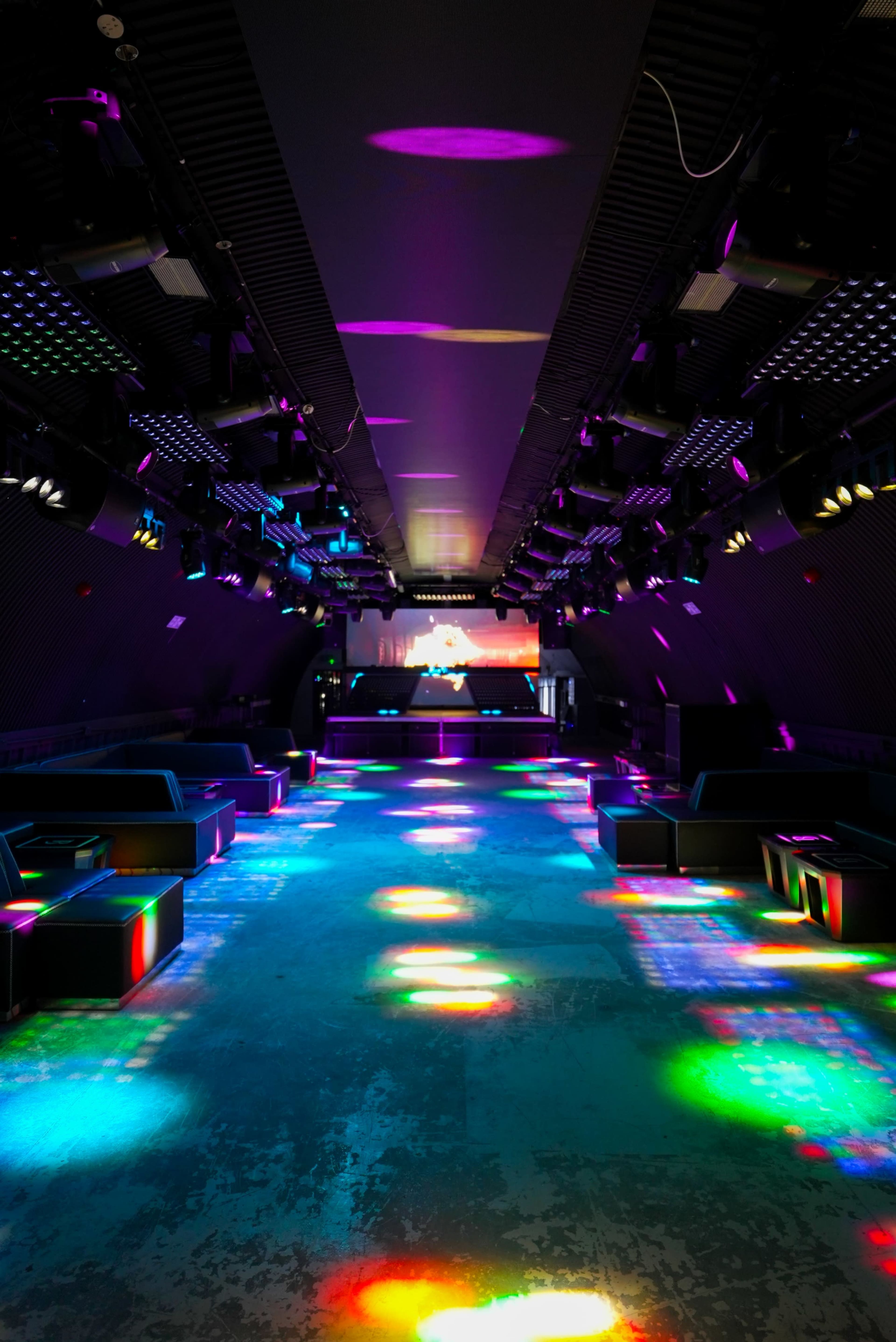 The image shows a spacious, dimly lit nightclub with colorful lights reflecting on the floor and seating areas.