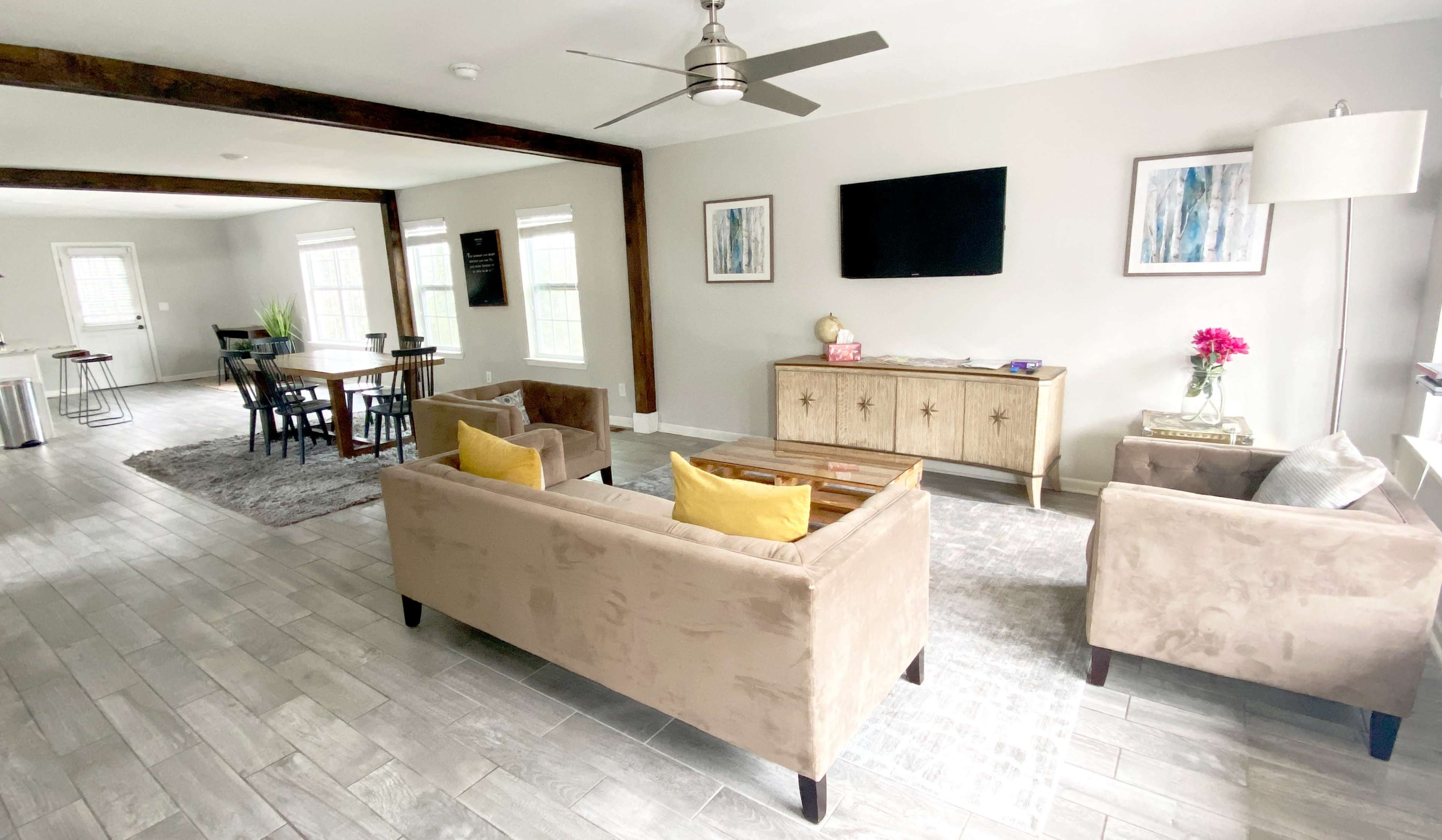 A modern living room features two beige sofas with yellow cushions, a wooden table, a television mounted on the wall, and large windows providing natural light.