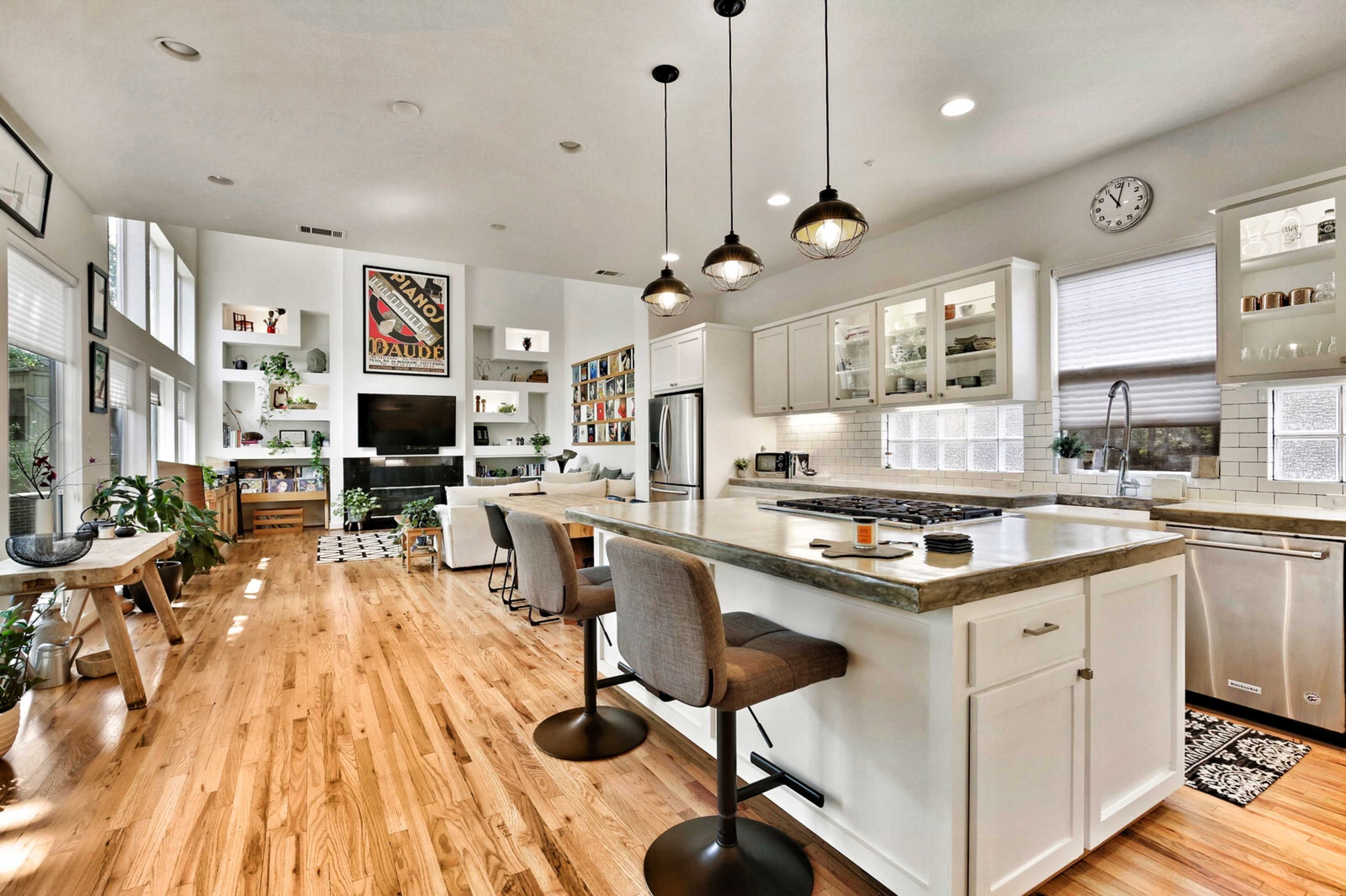 A modern kitchen features a central island with stools, white cabinets, wooden flooring, and a living area in the background with shelves and a television.