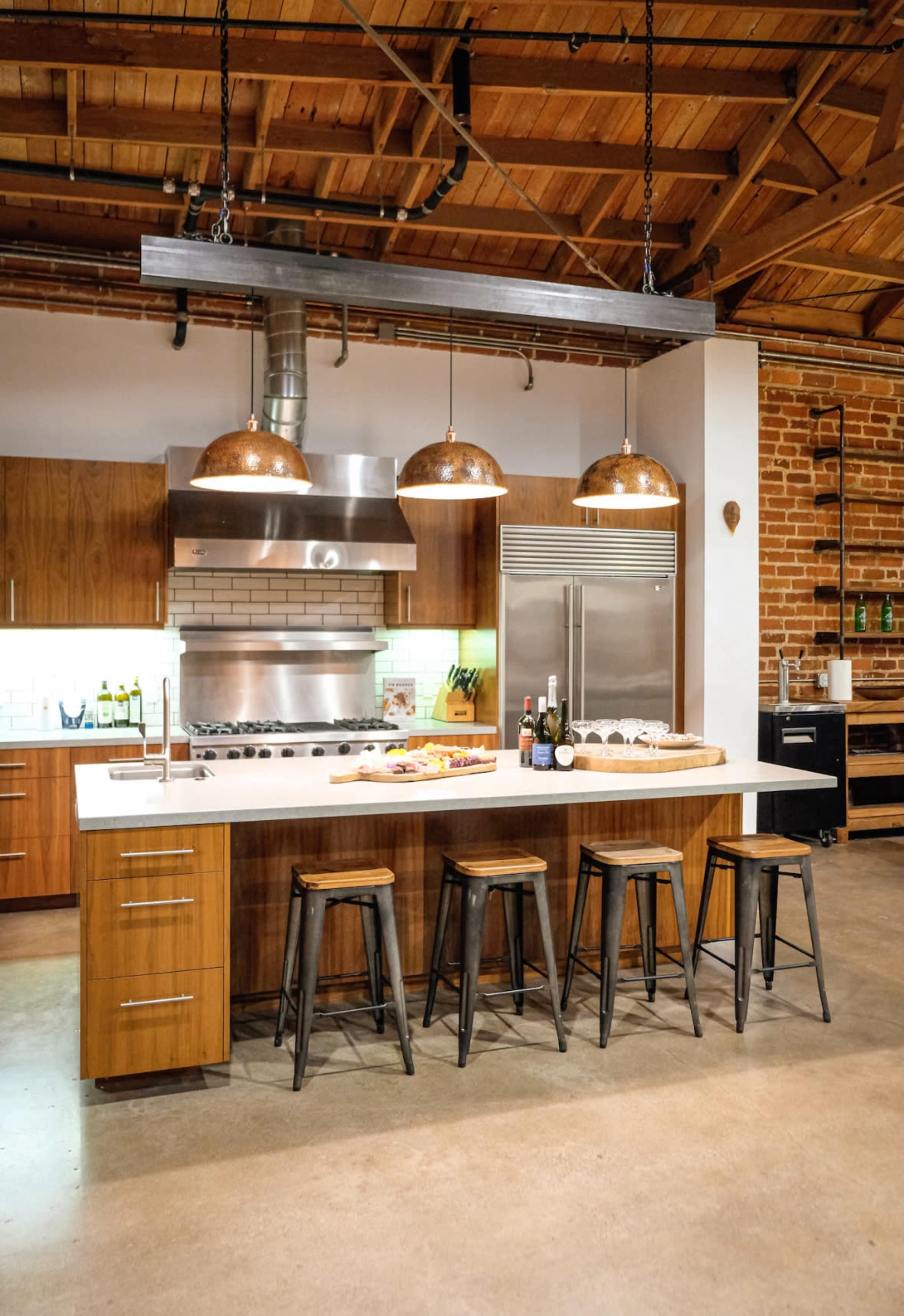 The image shows a modern kitchen featuring a central island with four metal stools, a stainless steel stove and refrigerator, and exposed wooden beams overhead.