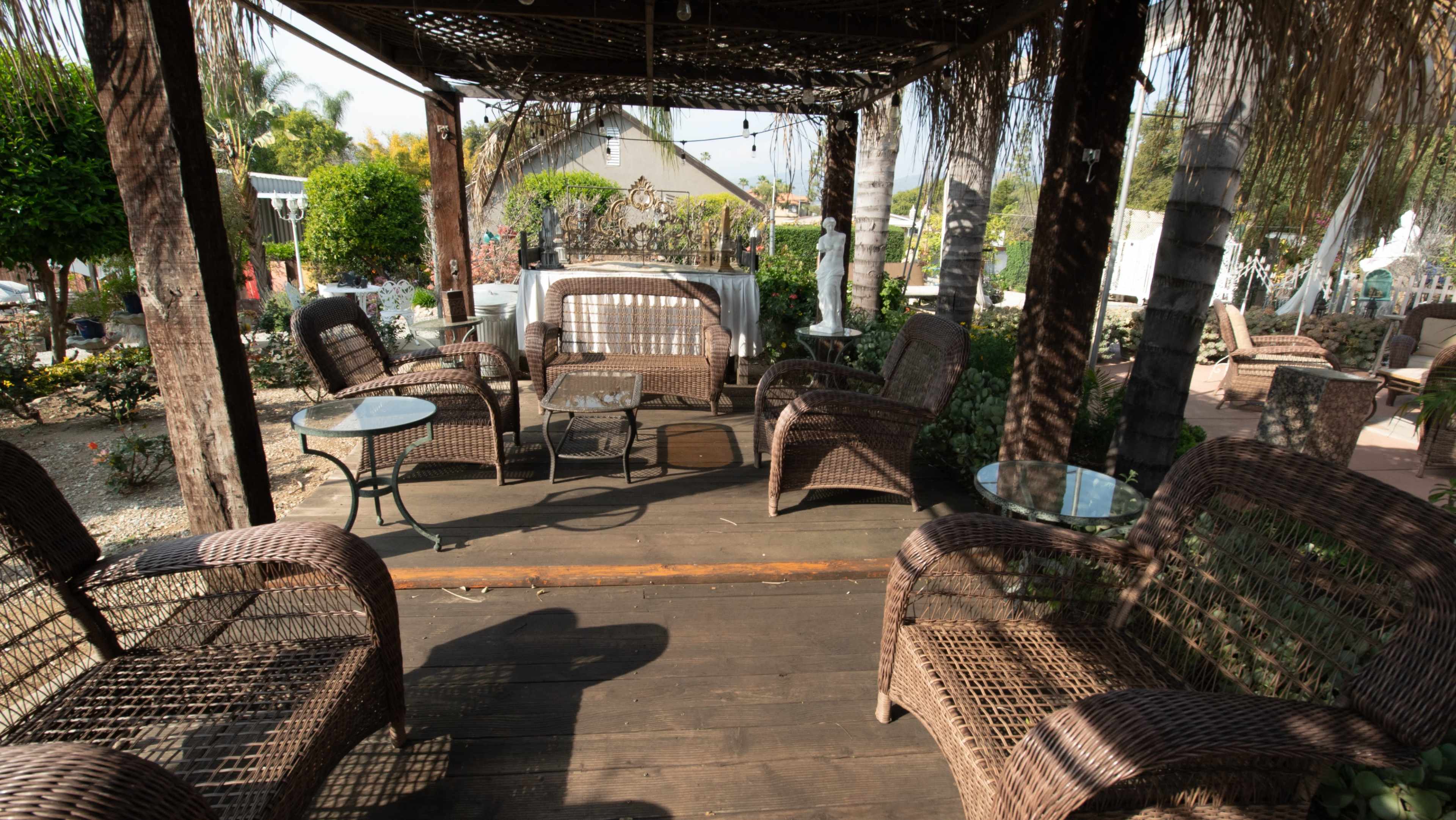 A shaded patio features several wicker chairs and glass-topped tables surrounded by greenery.