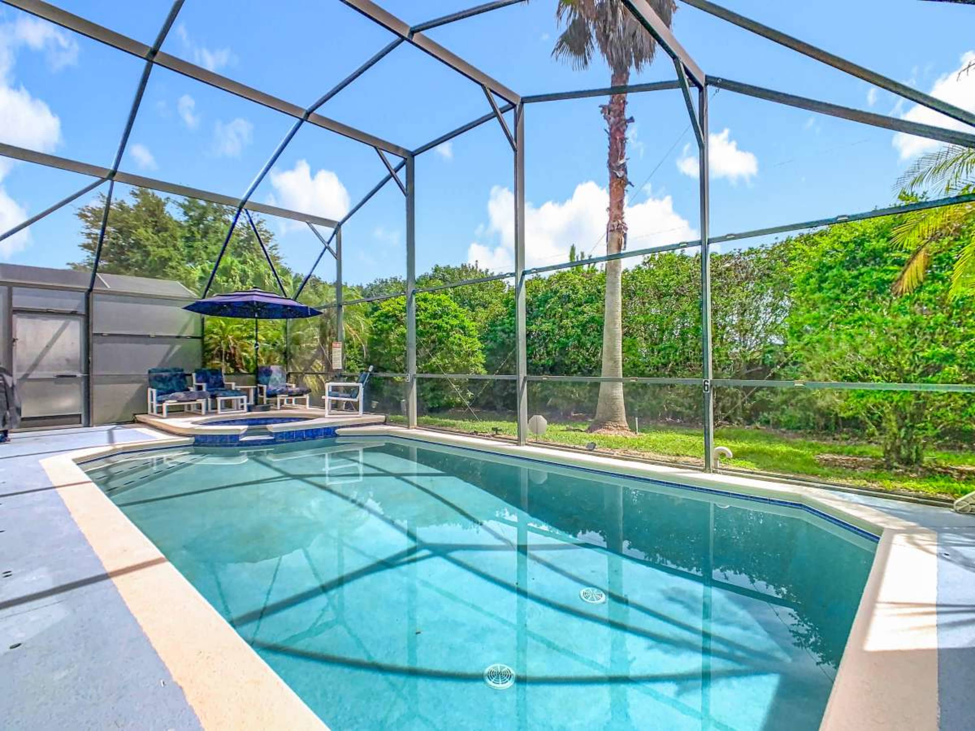 The image shows a screened-in pool area with a blue swimming pool, a lounge chair setup with an umbrella, and greenery in the background.