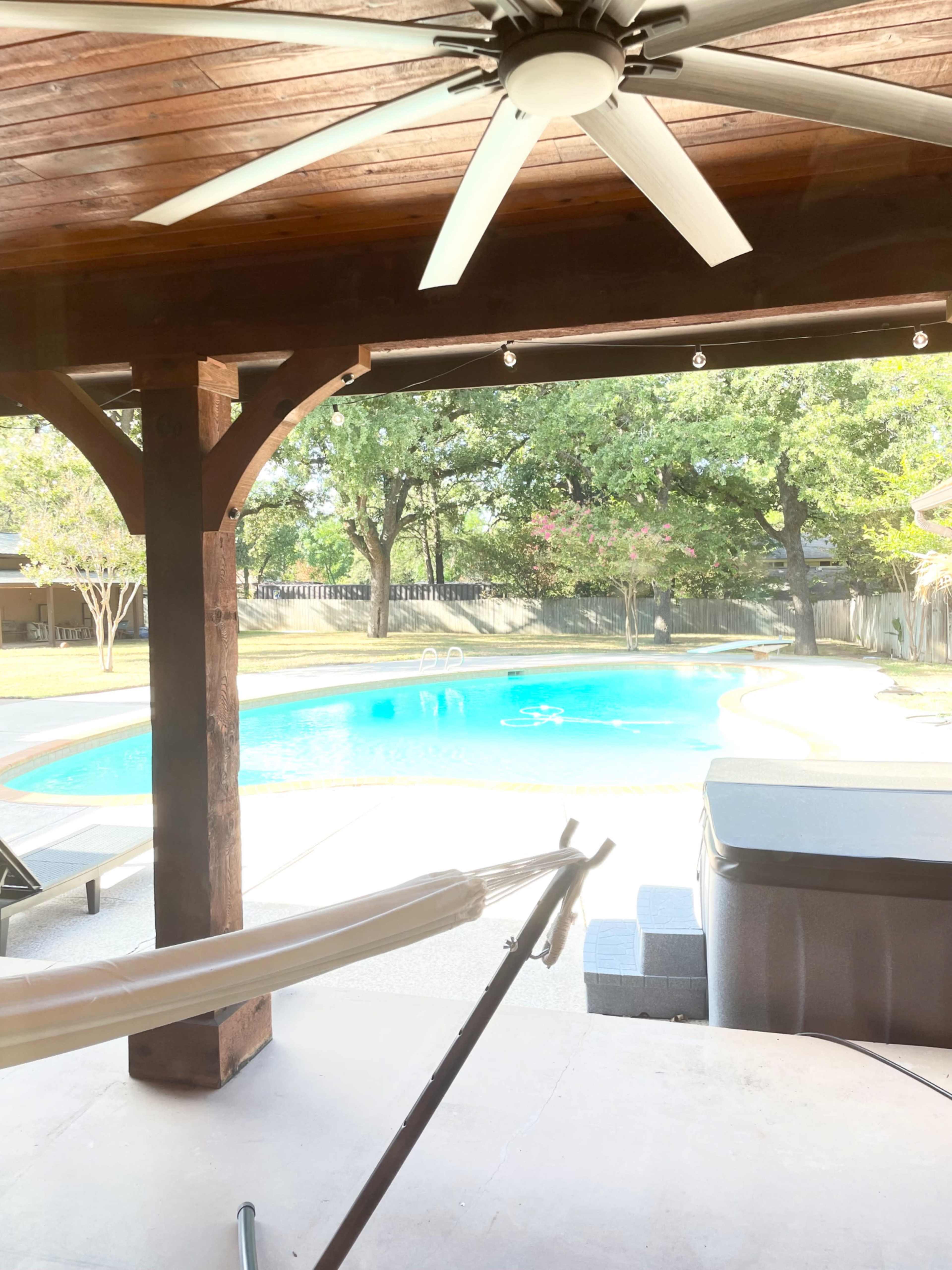 The image shows a covered patio with a ceiling fan overlooking a swimming pool surrounded by trees and outdoor furniture.