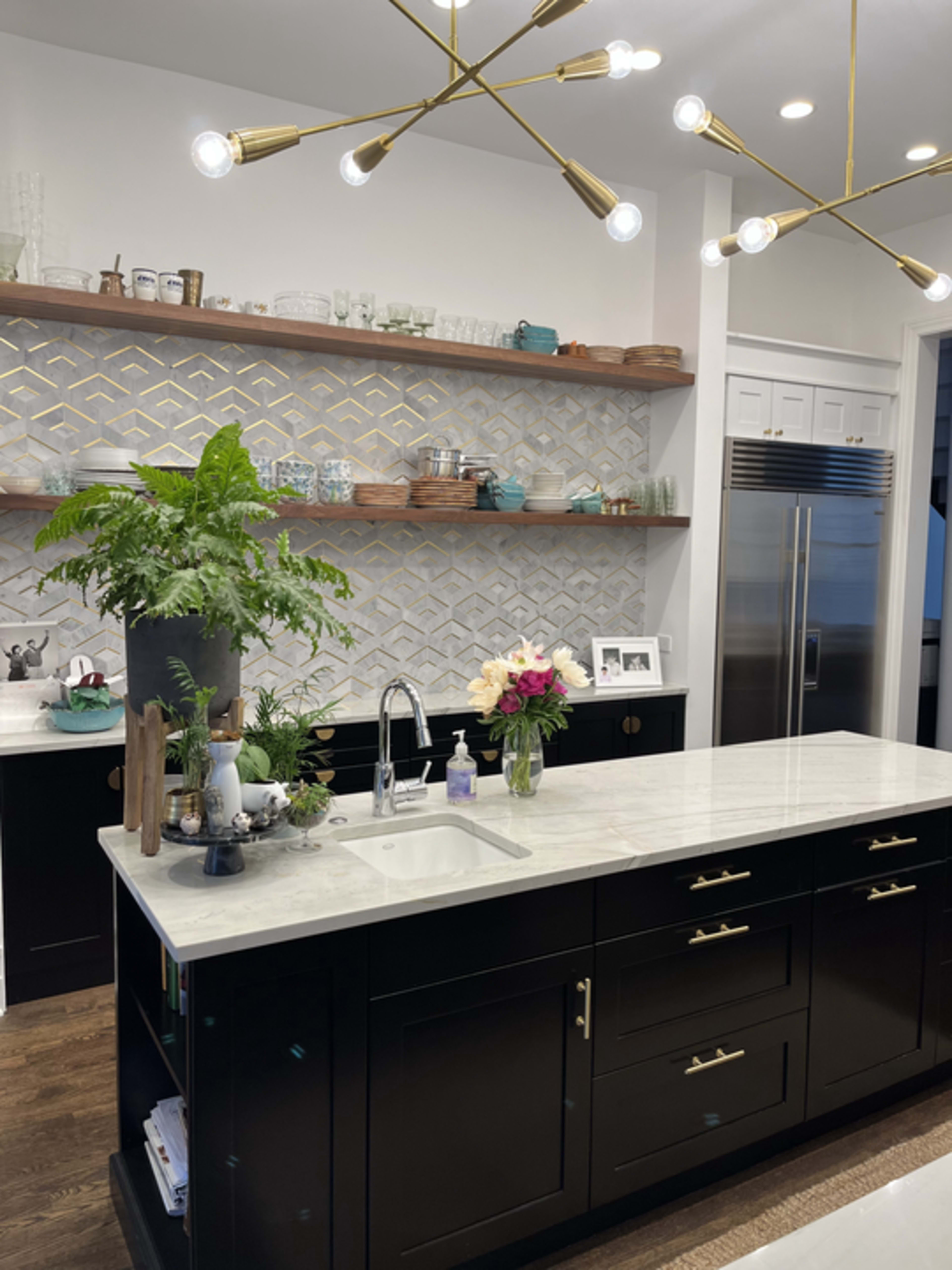 A modern kitchen features a large island with a marble countertop, black cabinetry, open shelves displaying glassware, and a decorative plant.