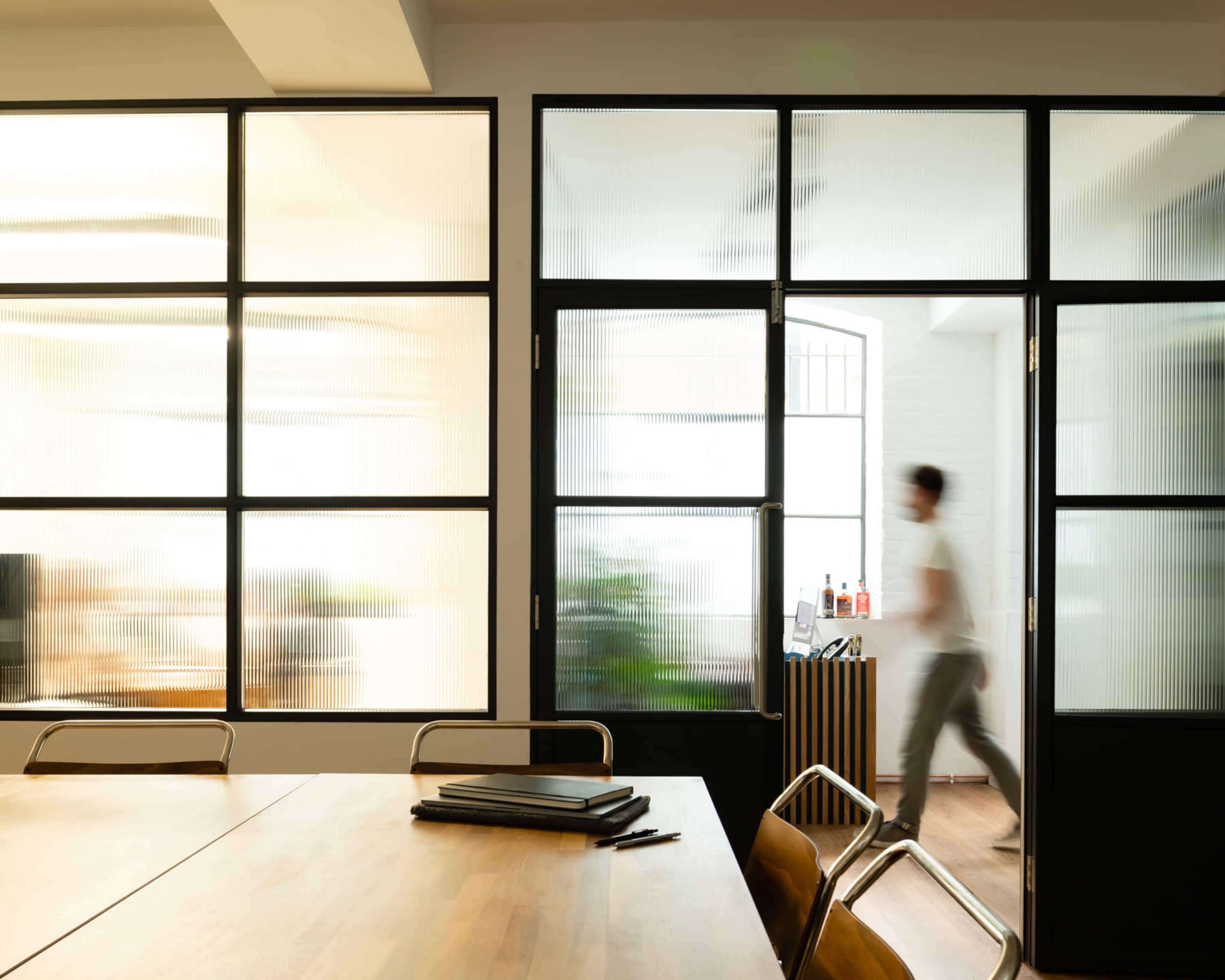 A person walks past frosted glass panels in an office, with a table and chairs visible in the foreground.