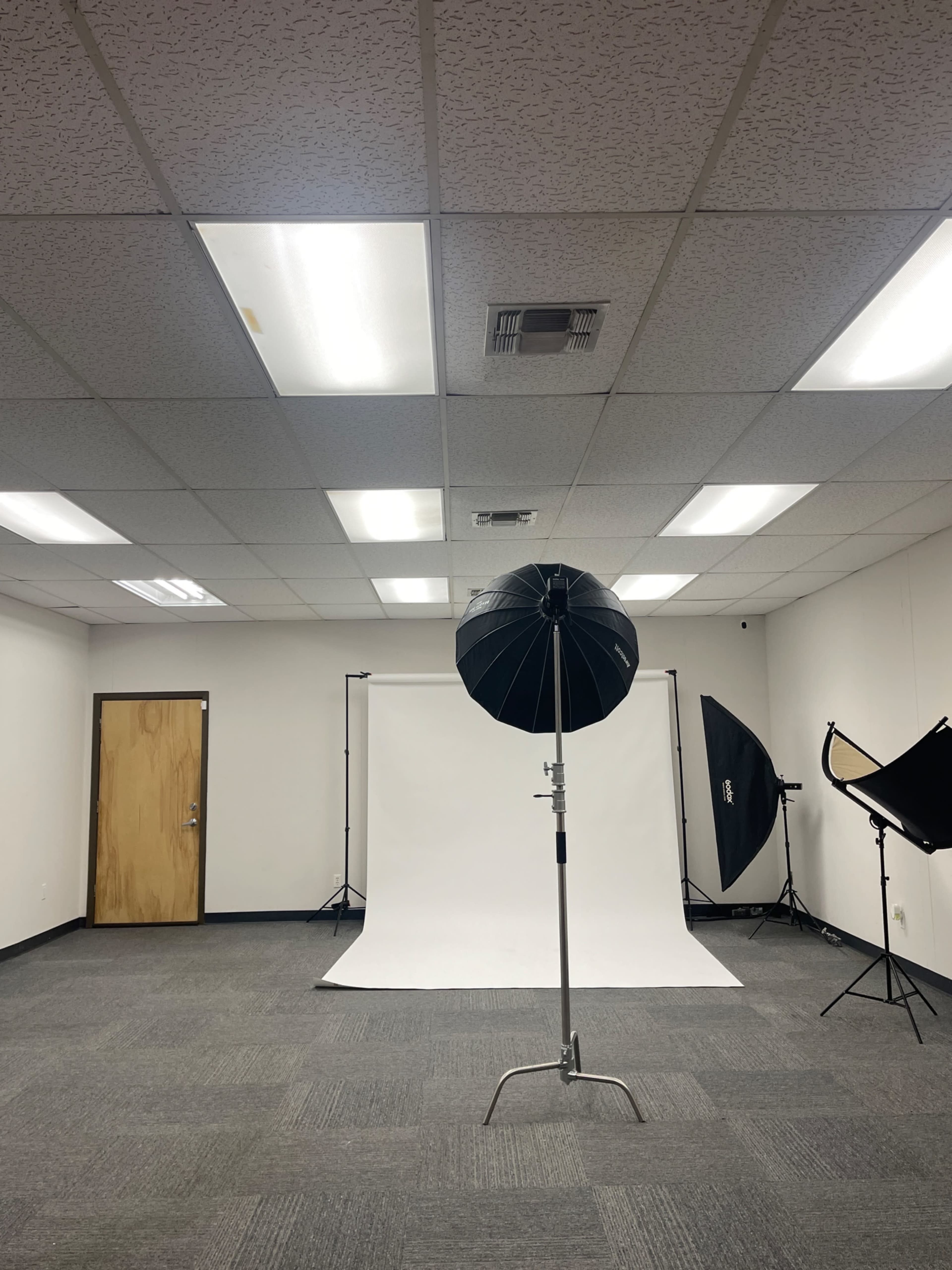 The image shows a photography studio with a white backdrop, lighting equipment, and a door on one side.