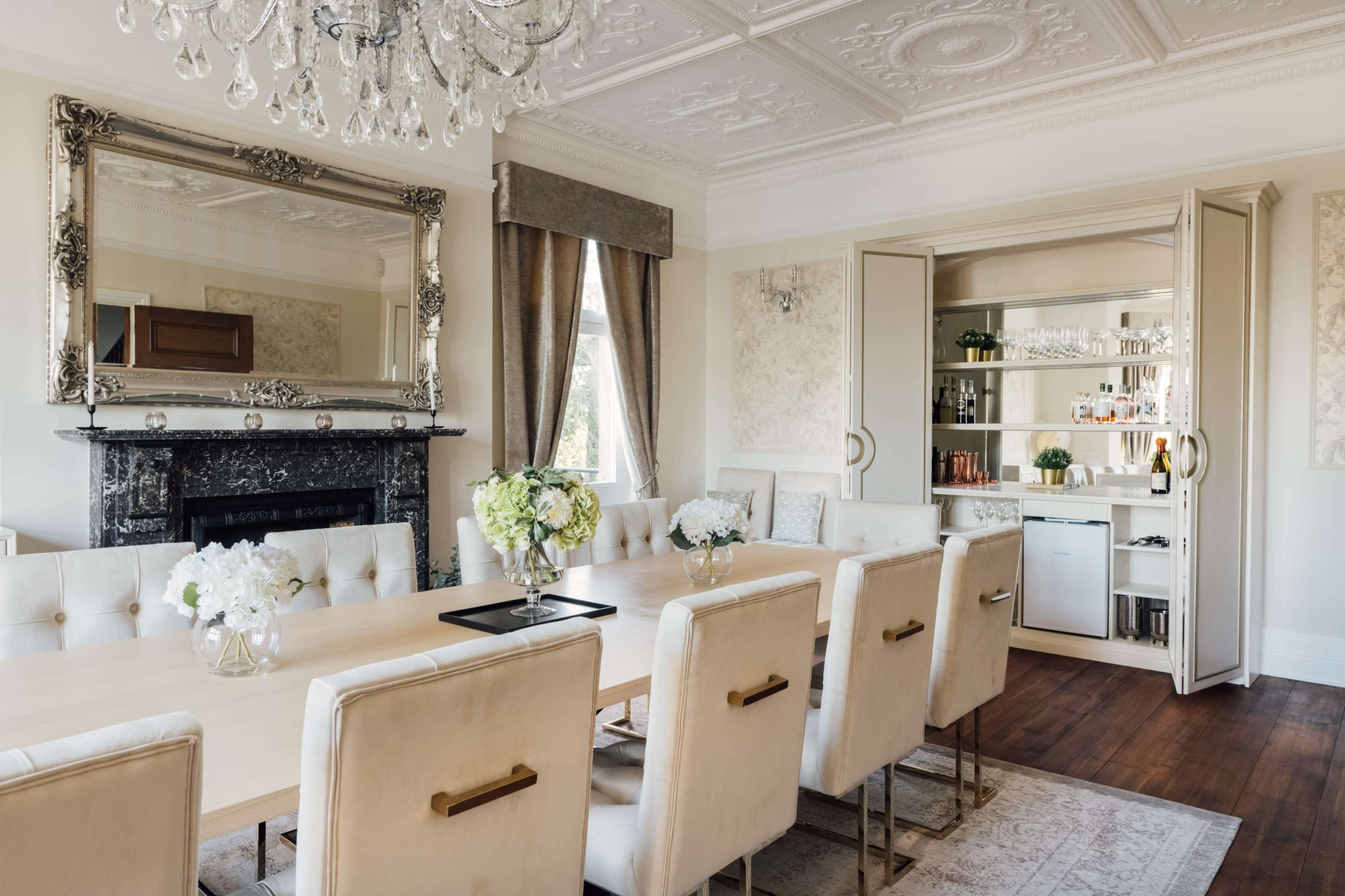 A formal dining room featuring a long wooden table surrounded by upholstered chairs, with a chandelier and a mirrored cabinet displaying glassware in the background.