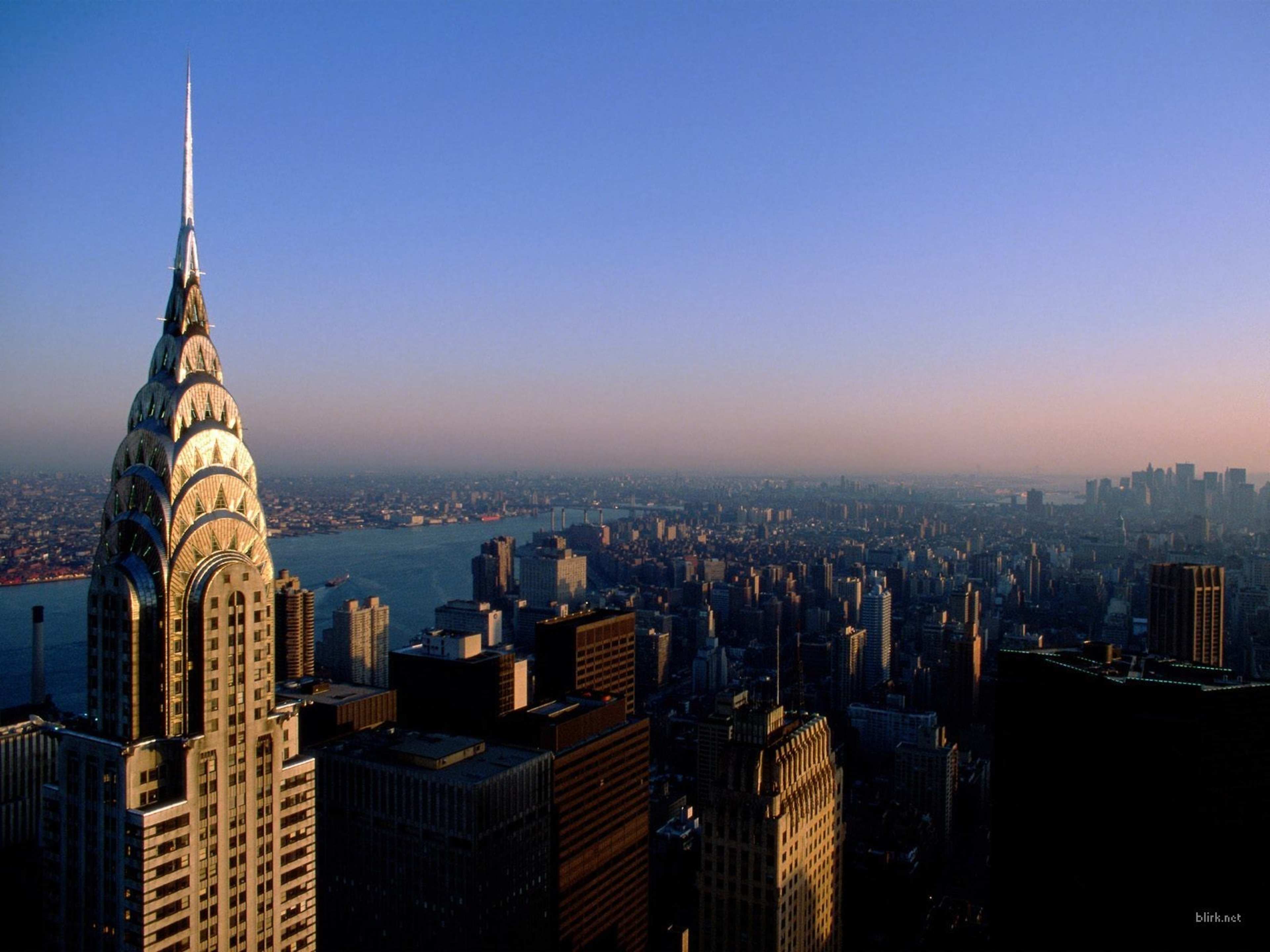 The image shows the spire of the Chrysler Building overlooking a city skyline at dusk with a body of water in the background.