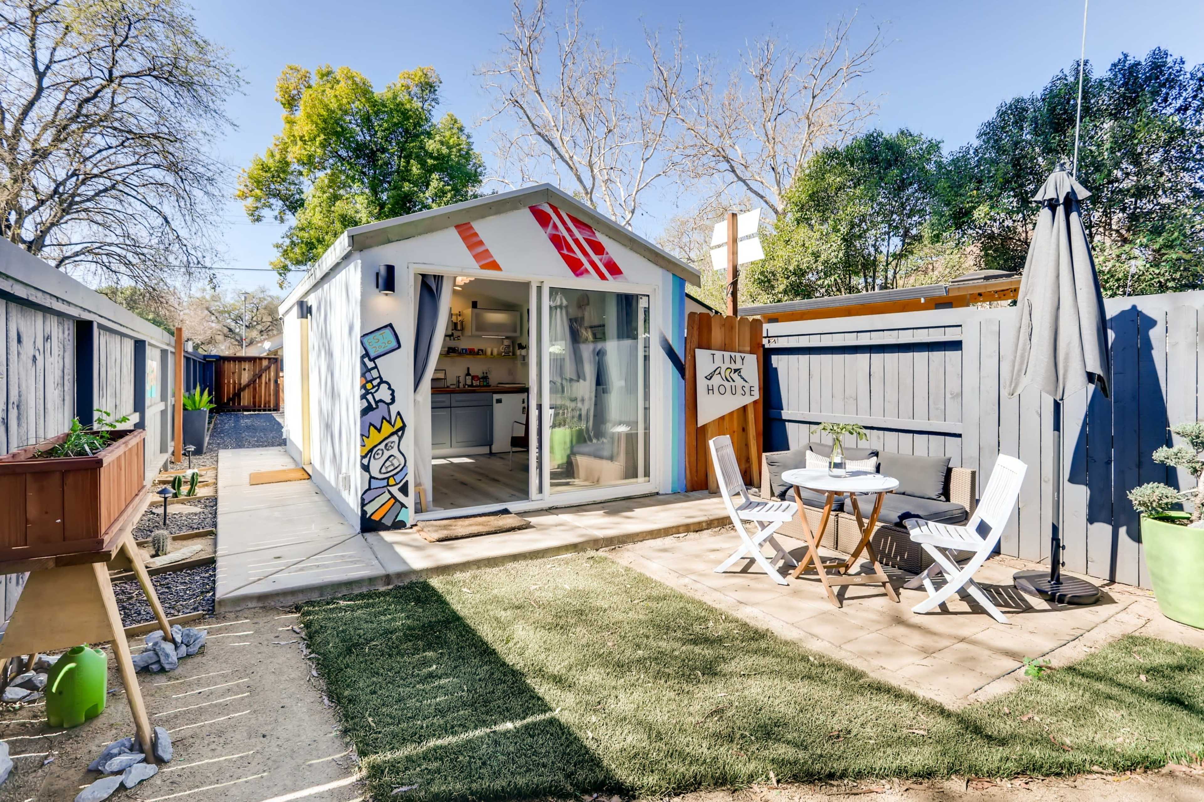 A small, modern shed-like structure with a bright mural and a patio featuring a table and chairs sits in a yard surrounded by greenery.