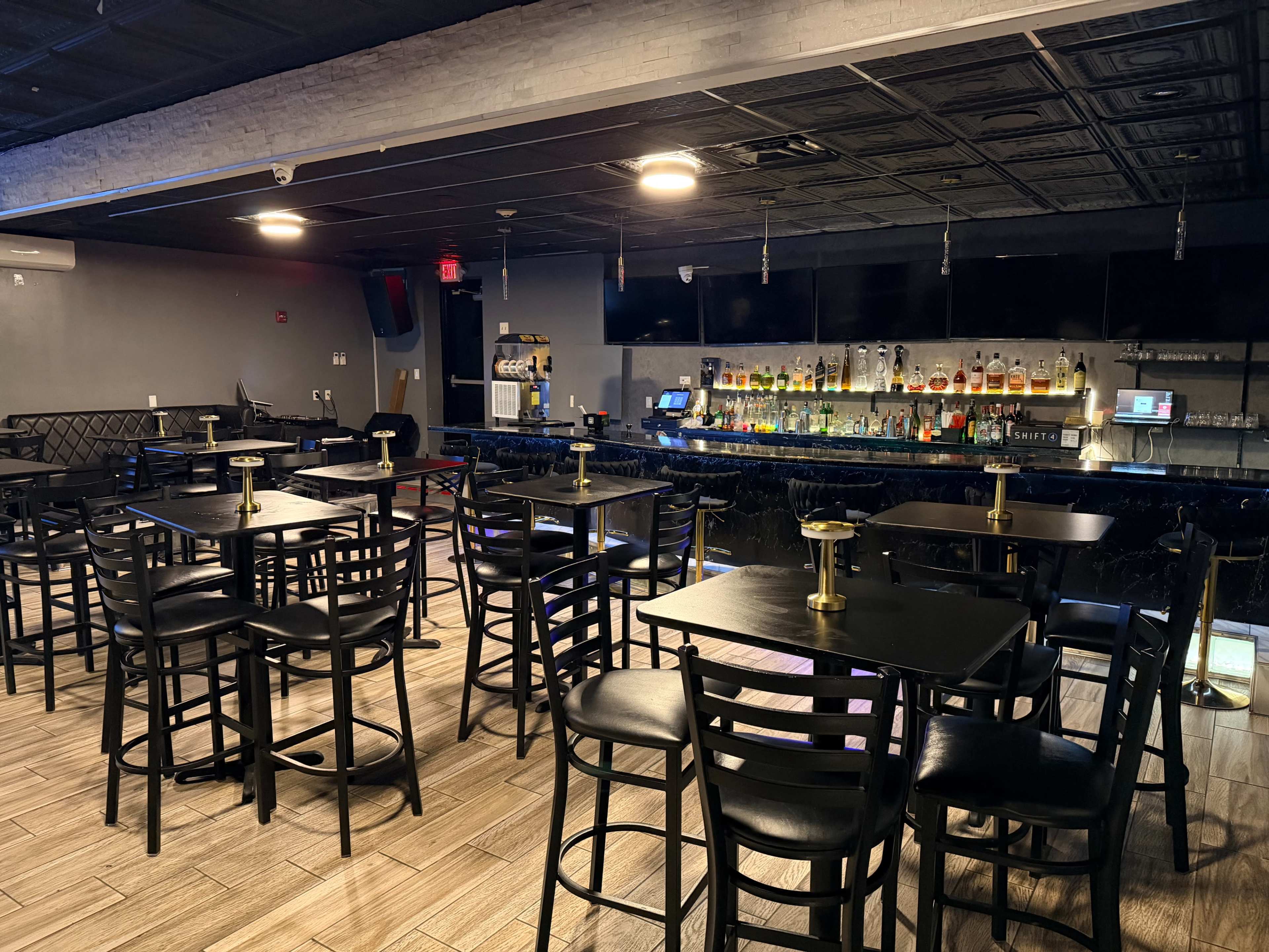 The image shows a bar interior with black tables and chairs, a well-stocked bar counter, and dim lighting.