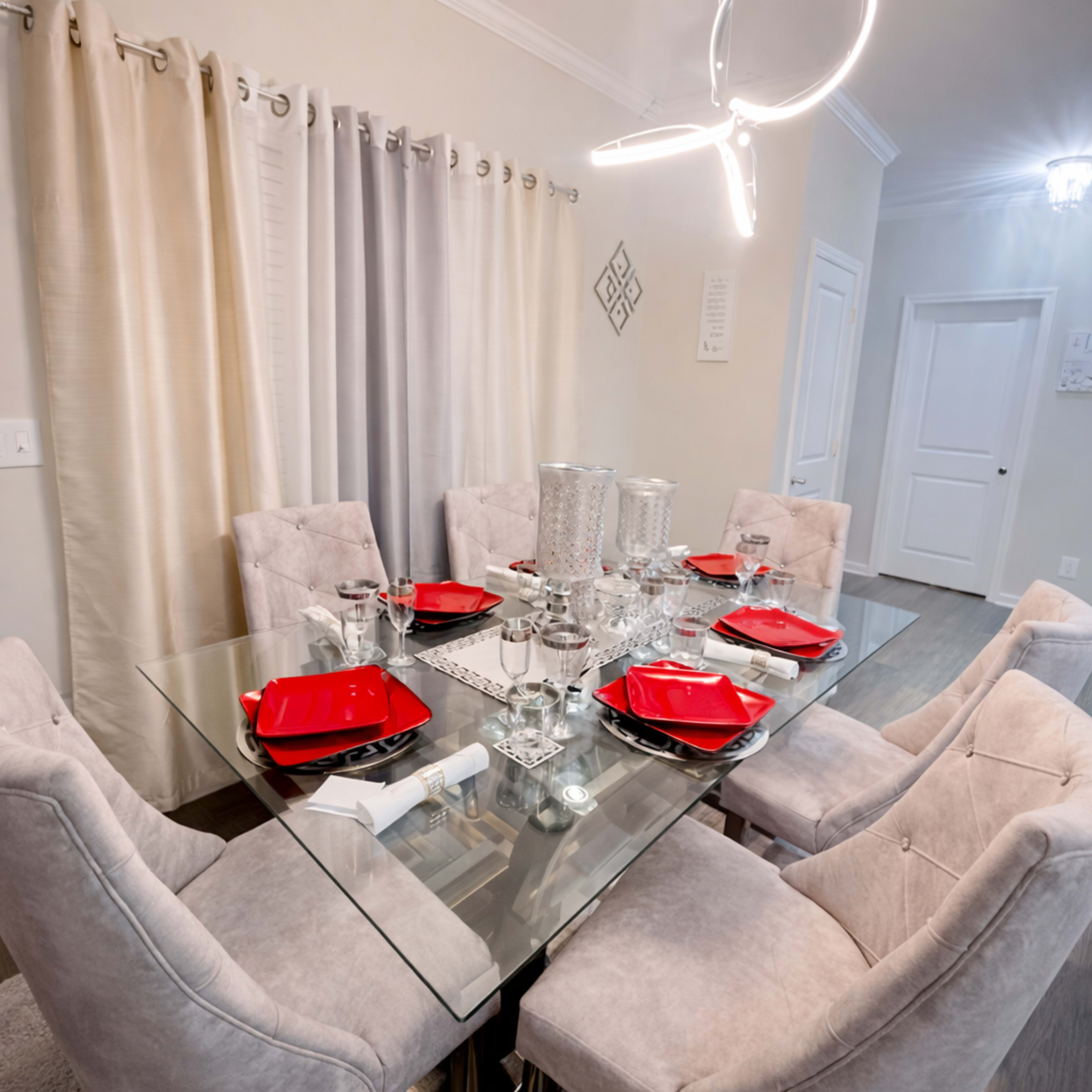 The image shows a modern dining table set for a meal, featuring a glass top, grey upholstered chairs, and red plates, with elegant tableware and decor.