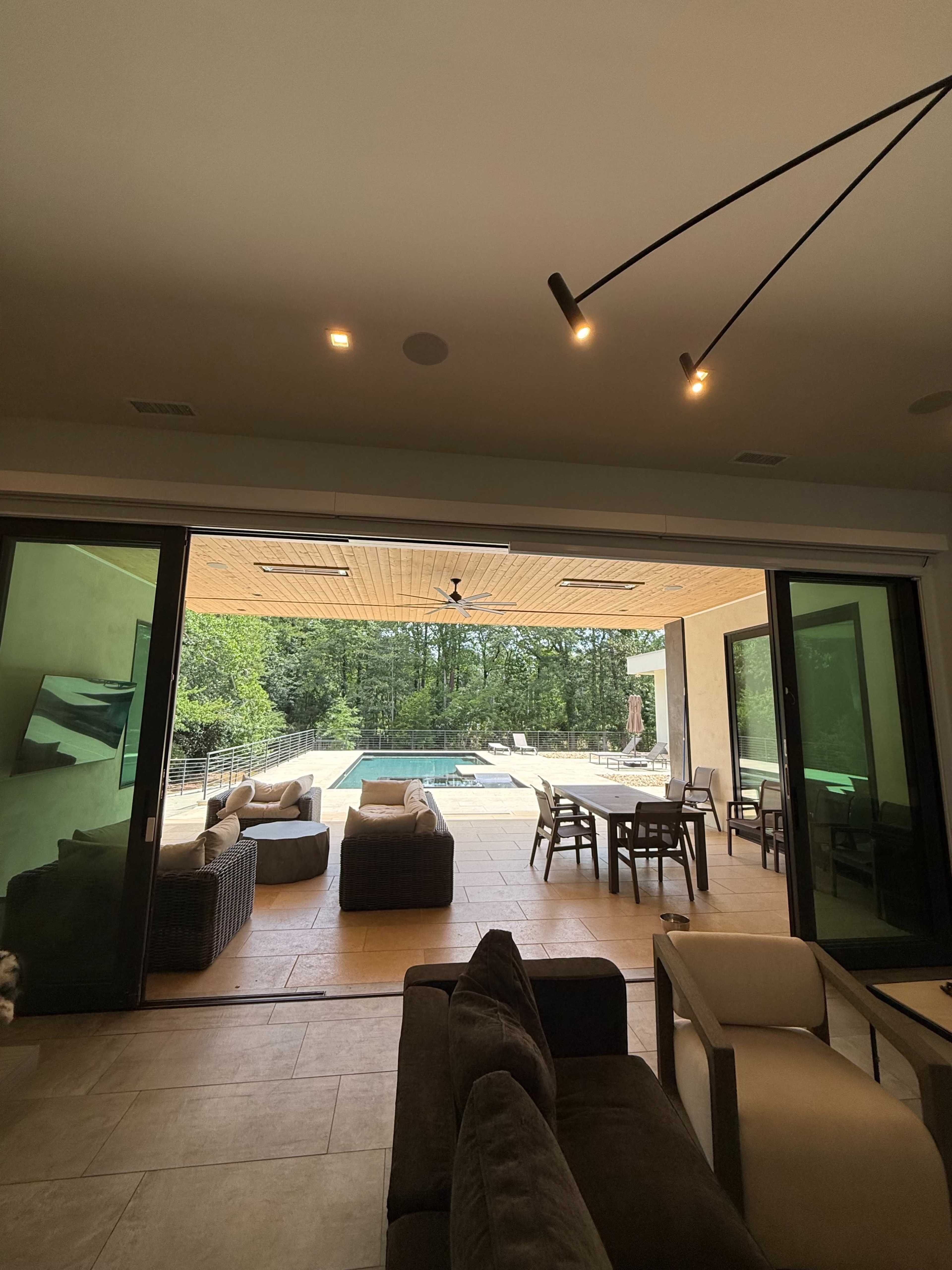 The image shows a modern indoor living area with large sliding glass doors leading to a patio that overlooks a swimming pool and wooded landscape.