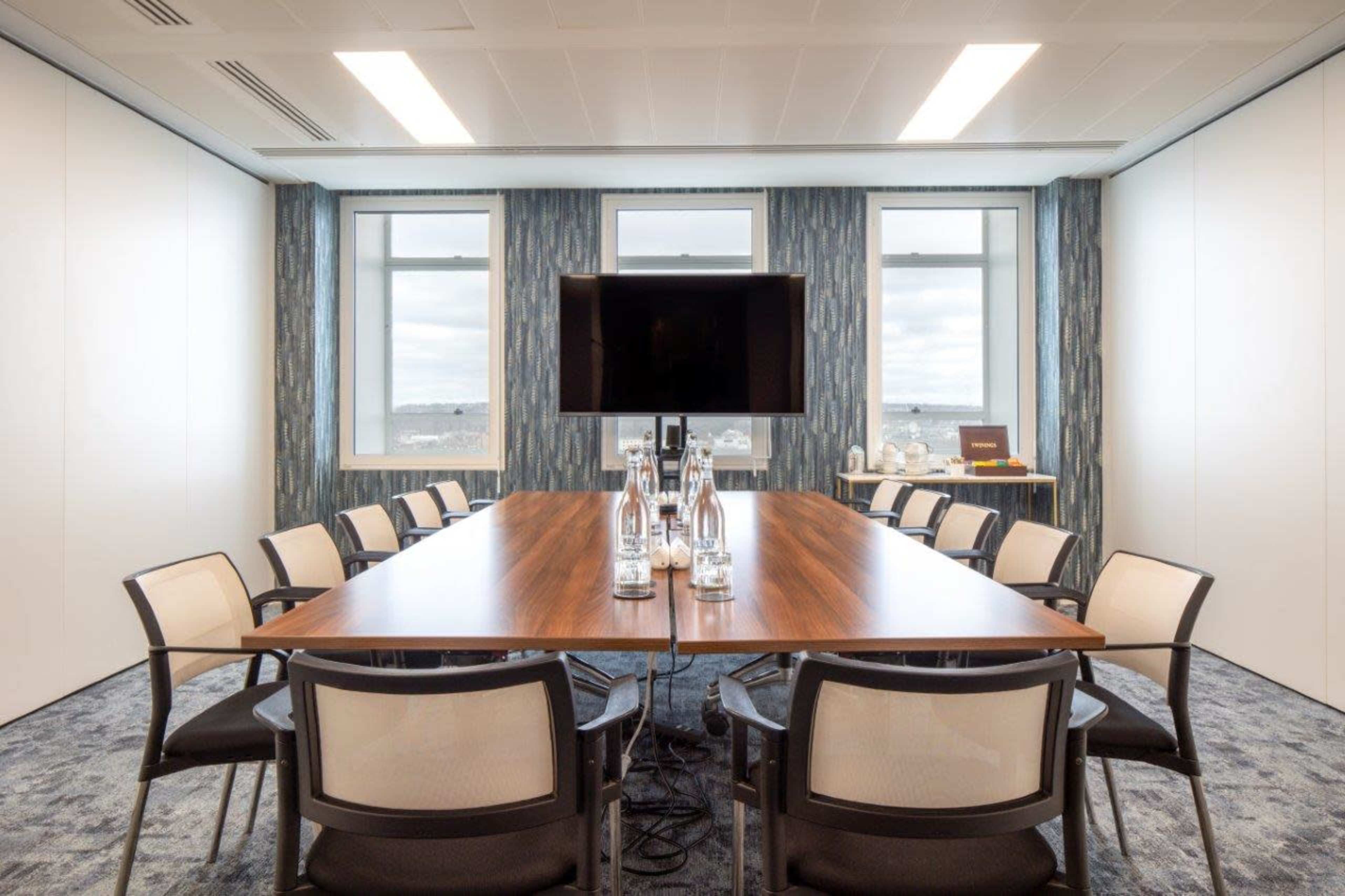 A modern conference room features a long wooden table surrounded by chairs, with a large monitor mounted on the wall and windows letting in natural light.