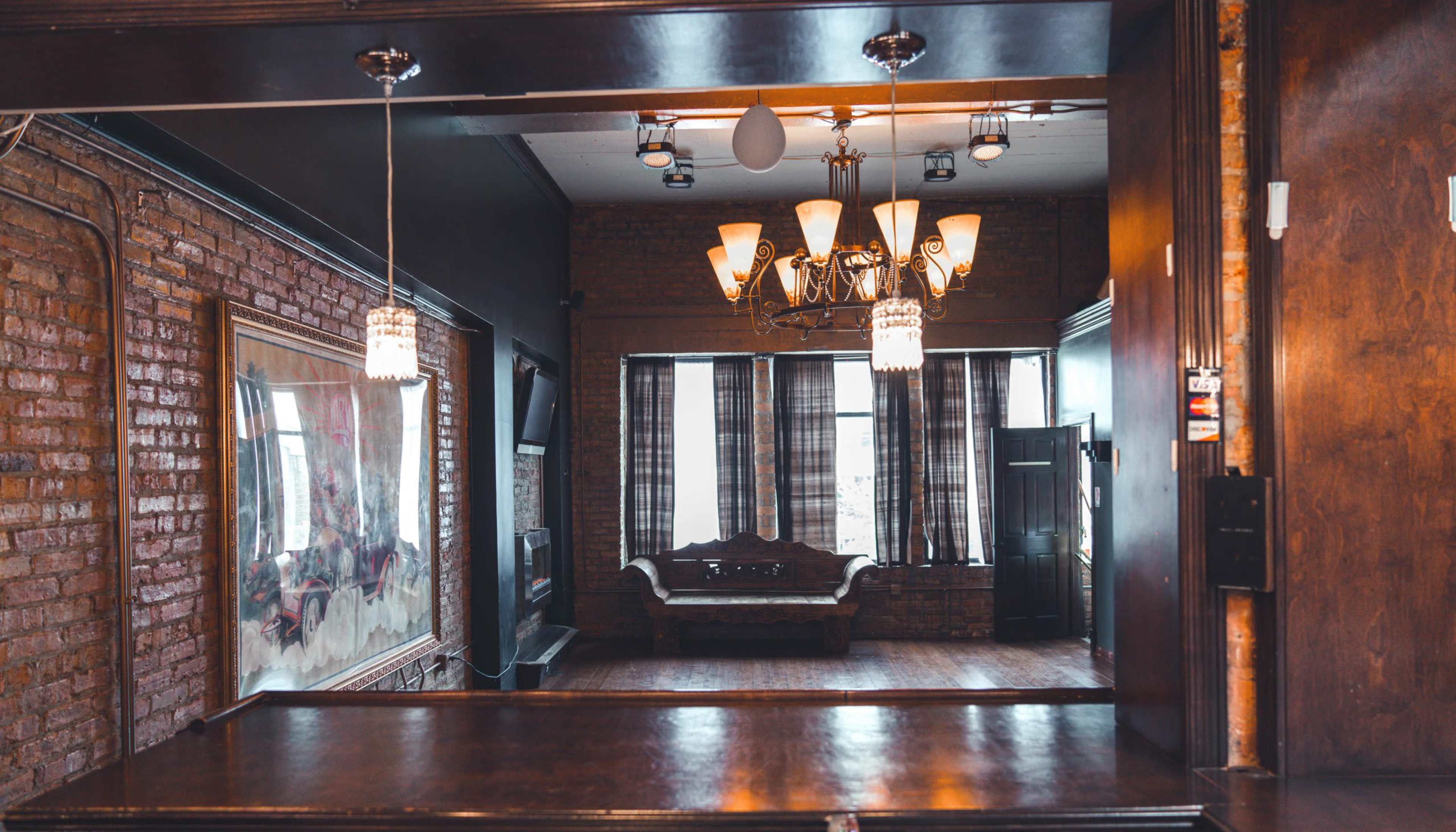 The image shows a spacious room featuring exposed brick walls, a chandelier, and a vintage couch near large windows with dark curtains.