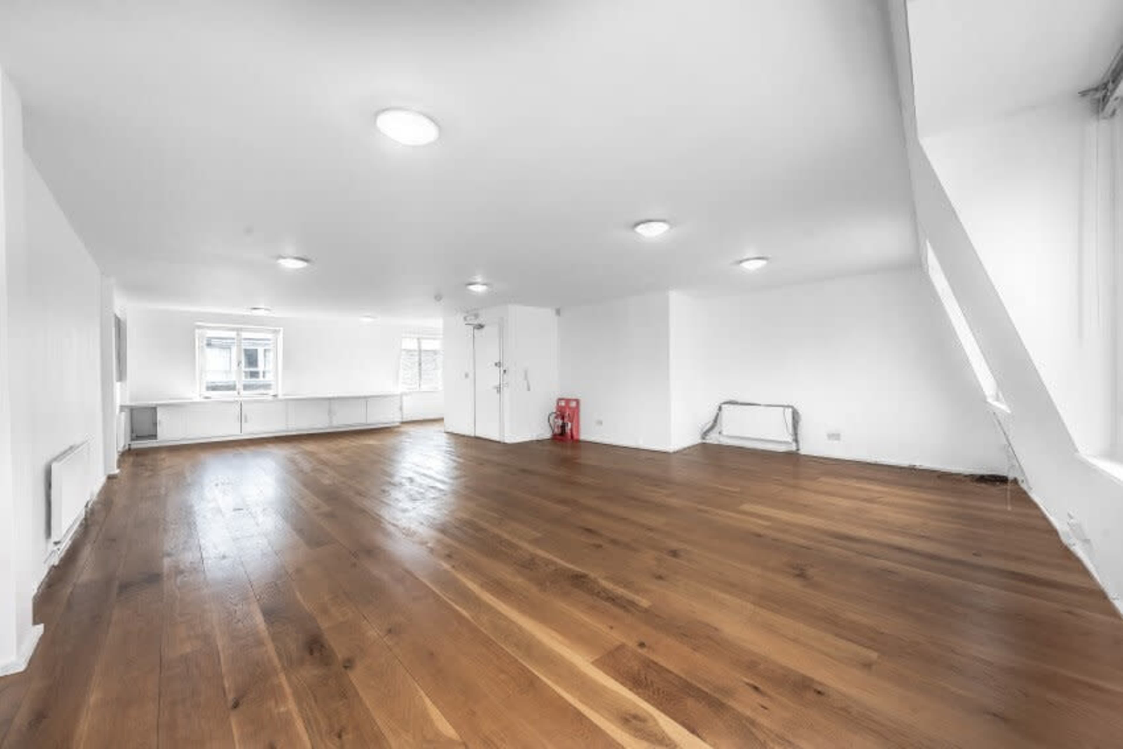 A spacious, empty room with wooden flooring and white walls, featuring several ceiling lights.