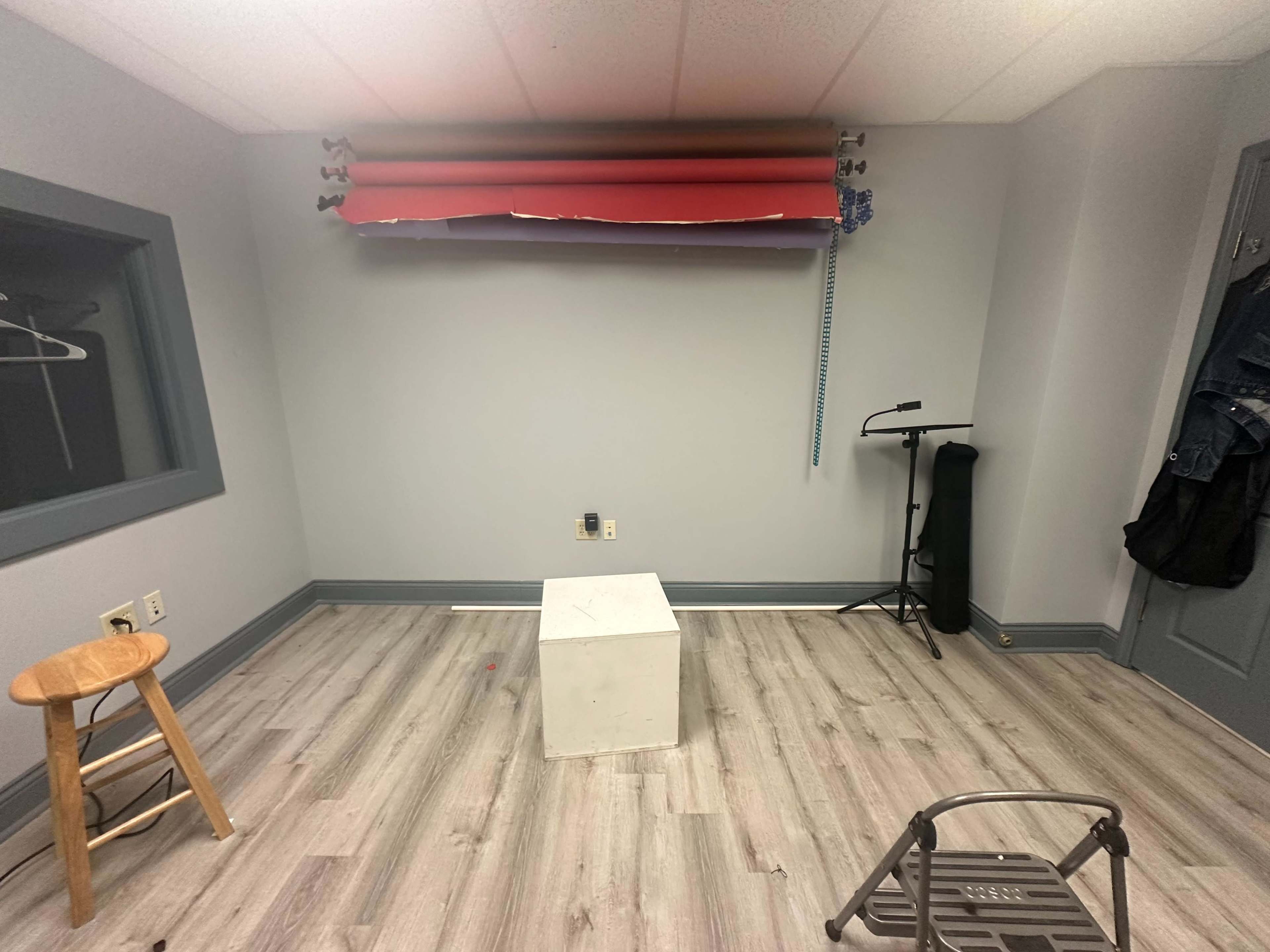 A sparse room with gray walls, a white cube in the center, a wooden stool, and a portable clothing rack.