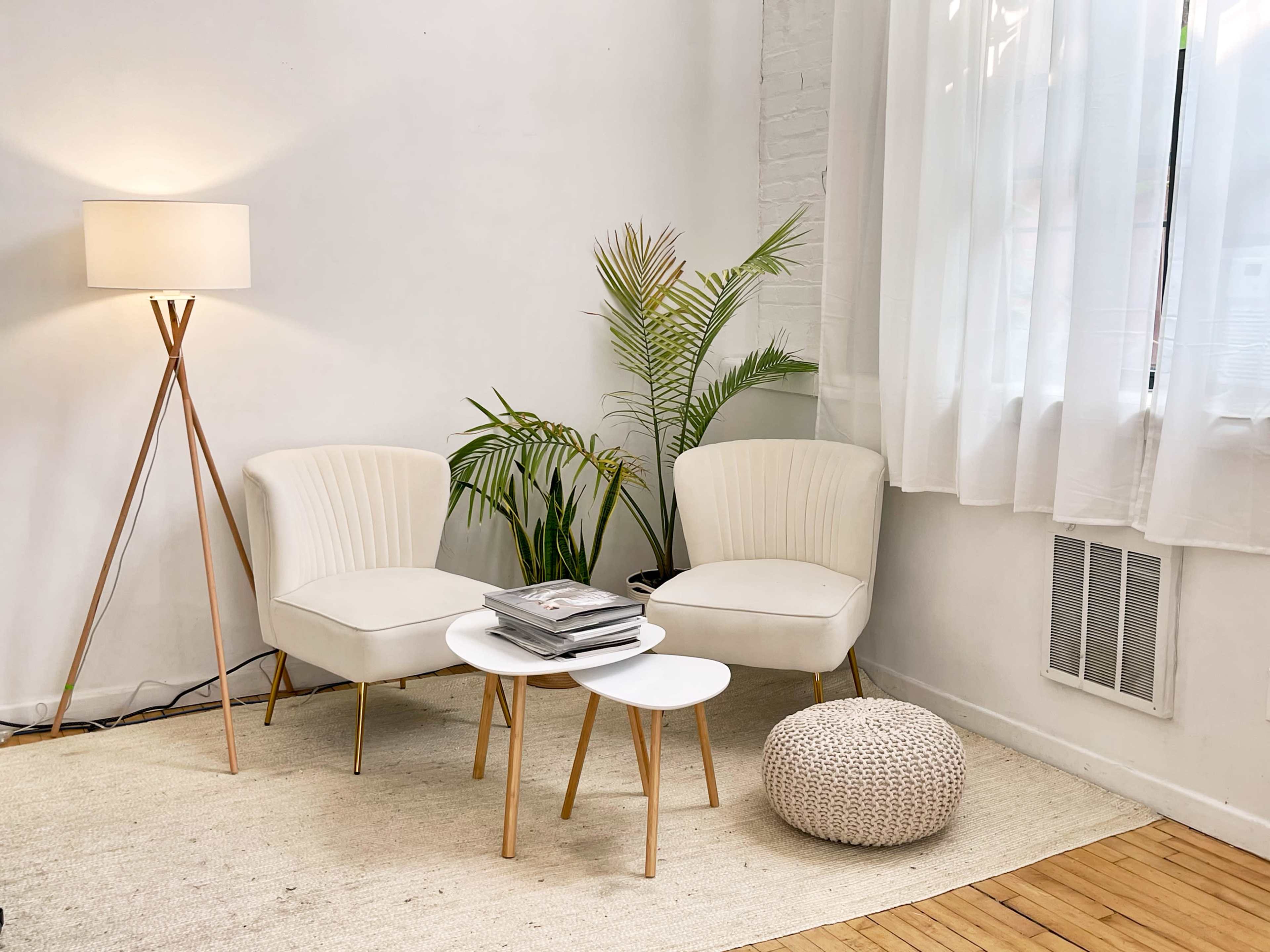 A bright, minimalist corner of a room with two white chairs, a small coffee table, a floor lamp, and a potted plant on a light rug.