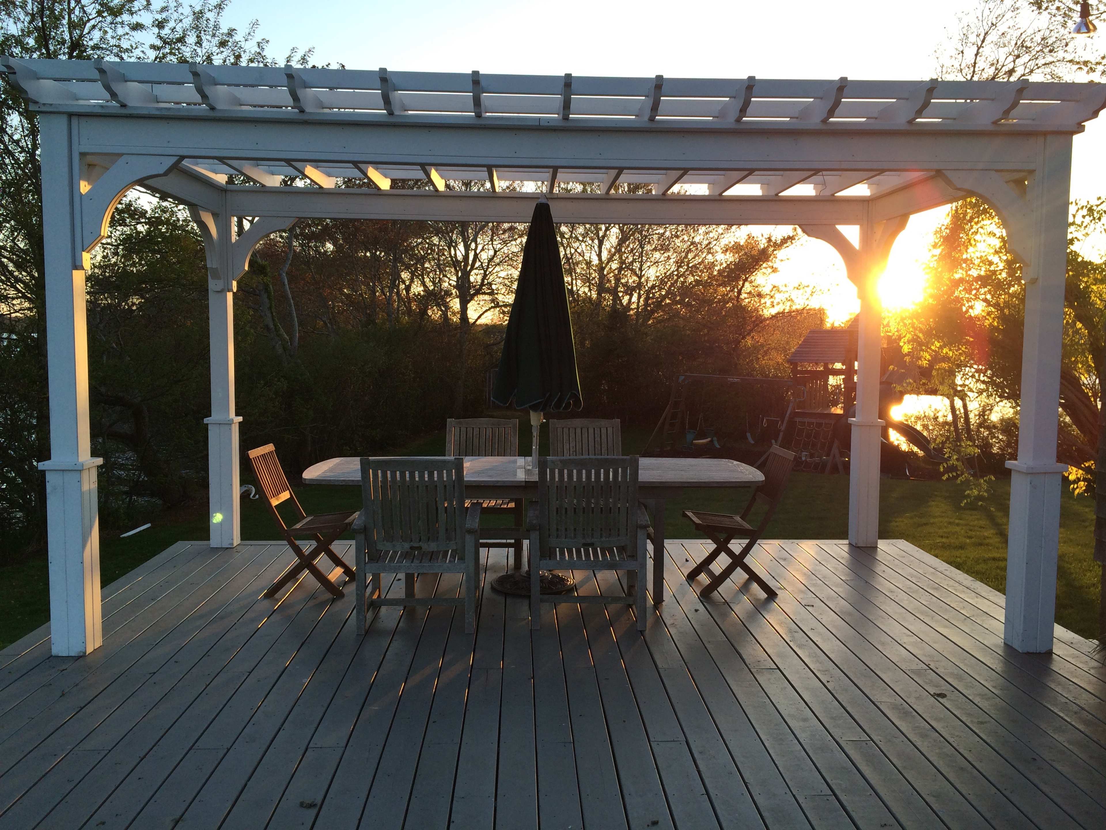 A white pergola with a shaded table and chairs sits on a wooden deck, framed by a sunset over a tranquil landscape.