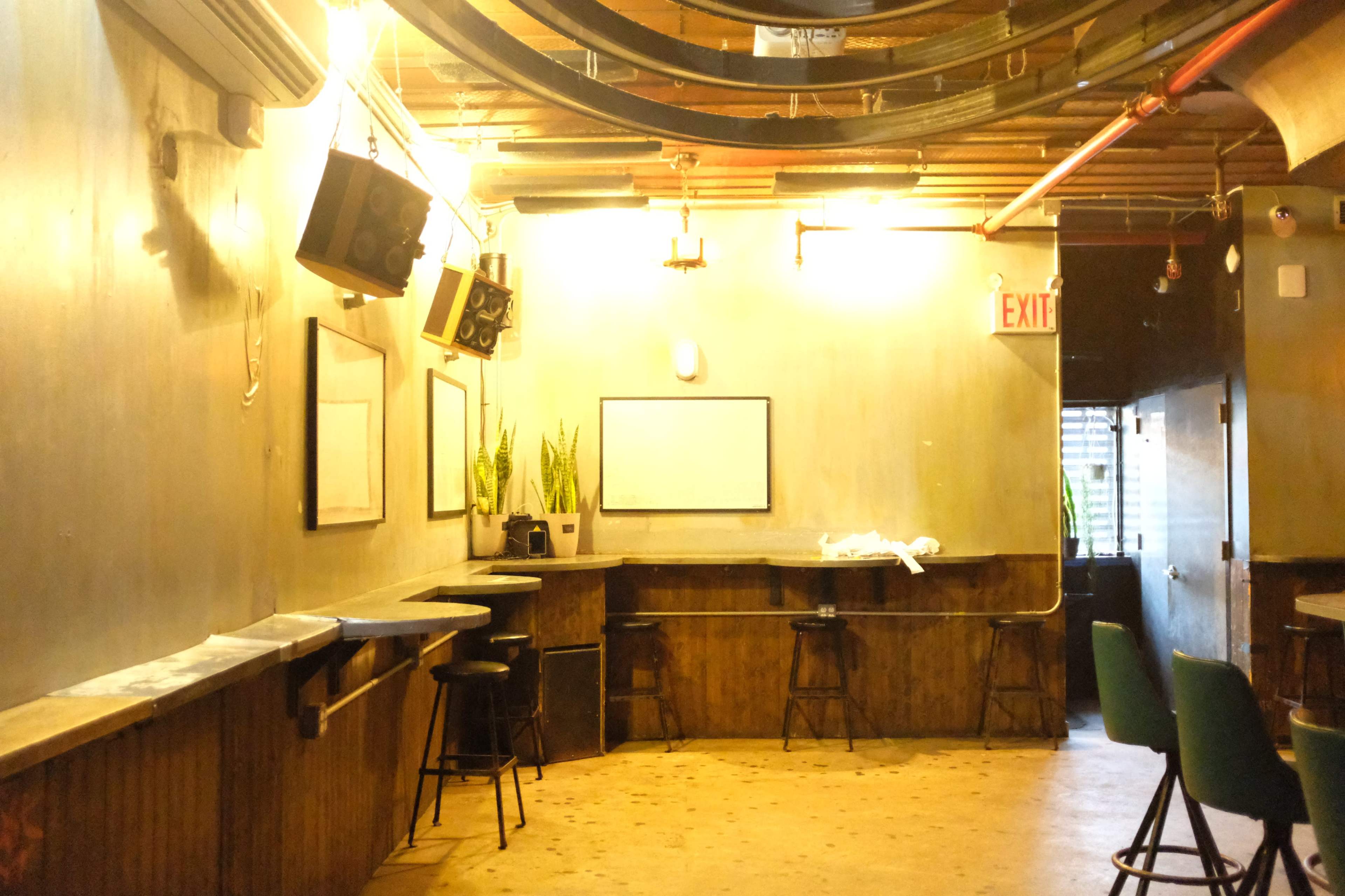 The image shows a sparsely furnished, dimly lit room featuring wooden bar stools and mirrors along the wall, with plants in the corner and an exit sign visible.