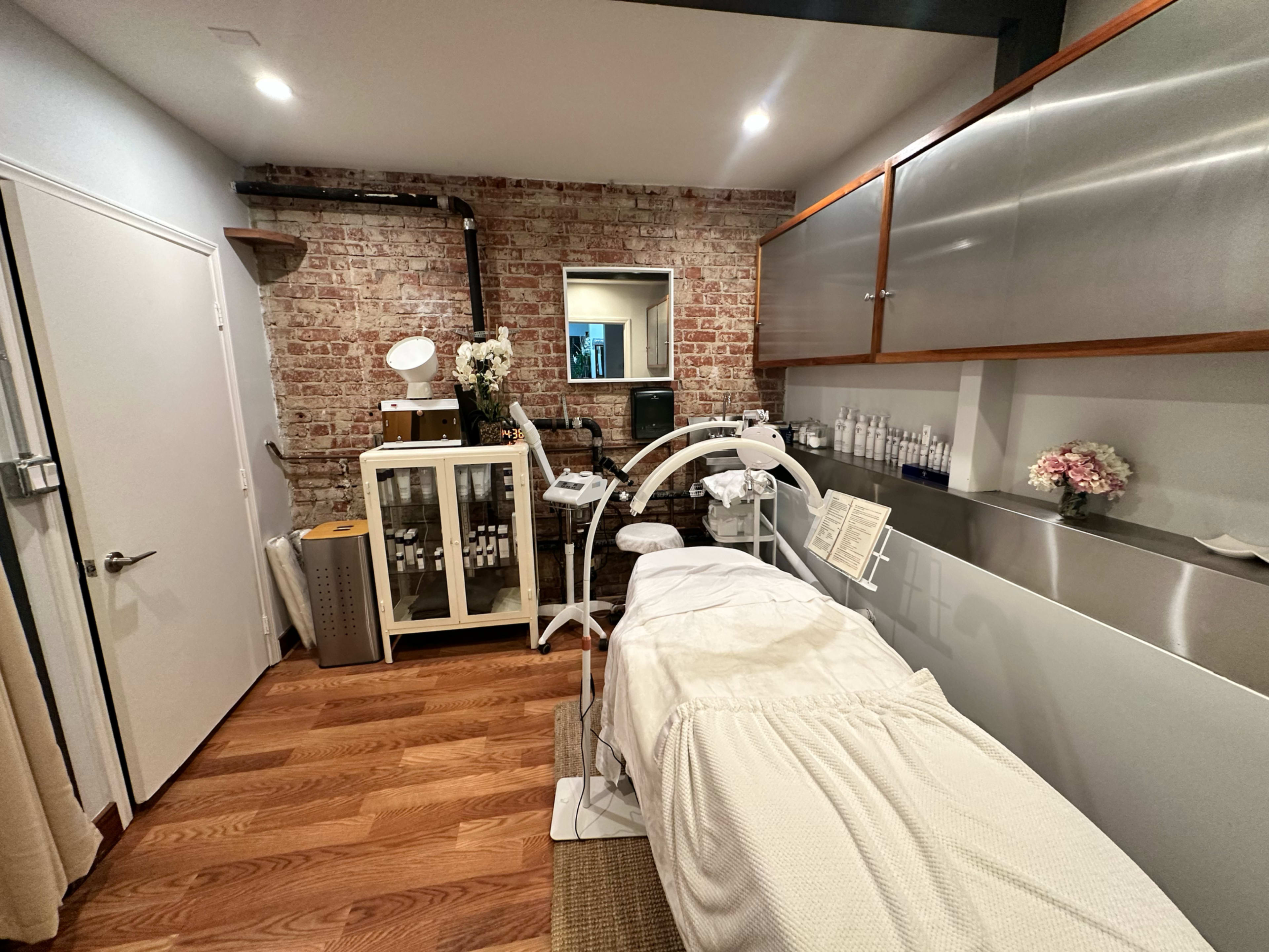 The image shows a clean, modern skincare treatment room with a treatment bed, storage units, and exposed brick walls.