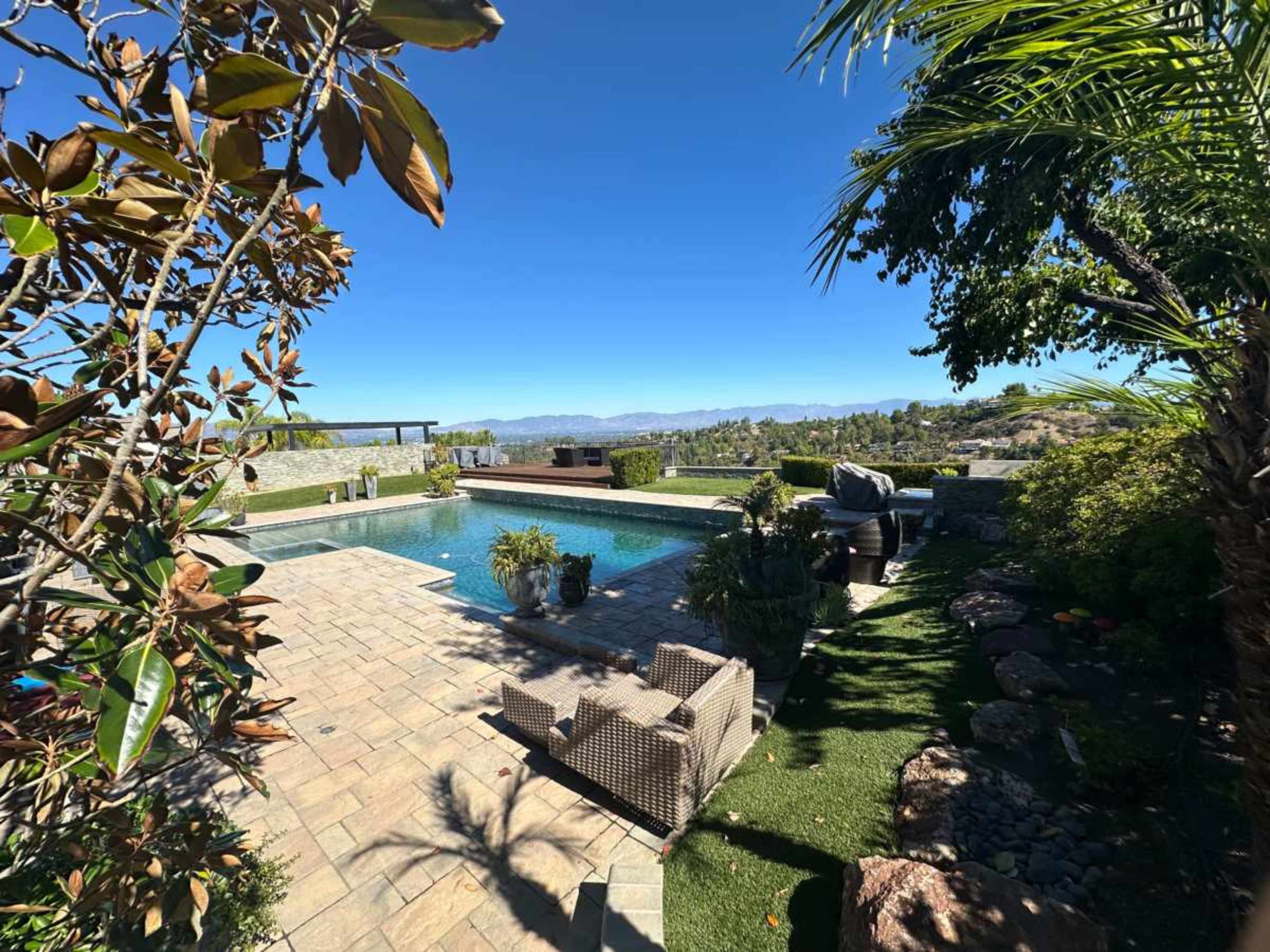 The image shows a landscaped backyard with a swimming pool, surrounded by stone pathways and various plants, under a clear blue sky.
