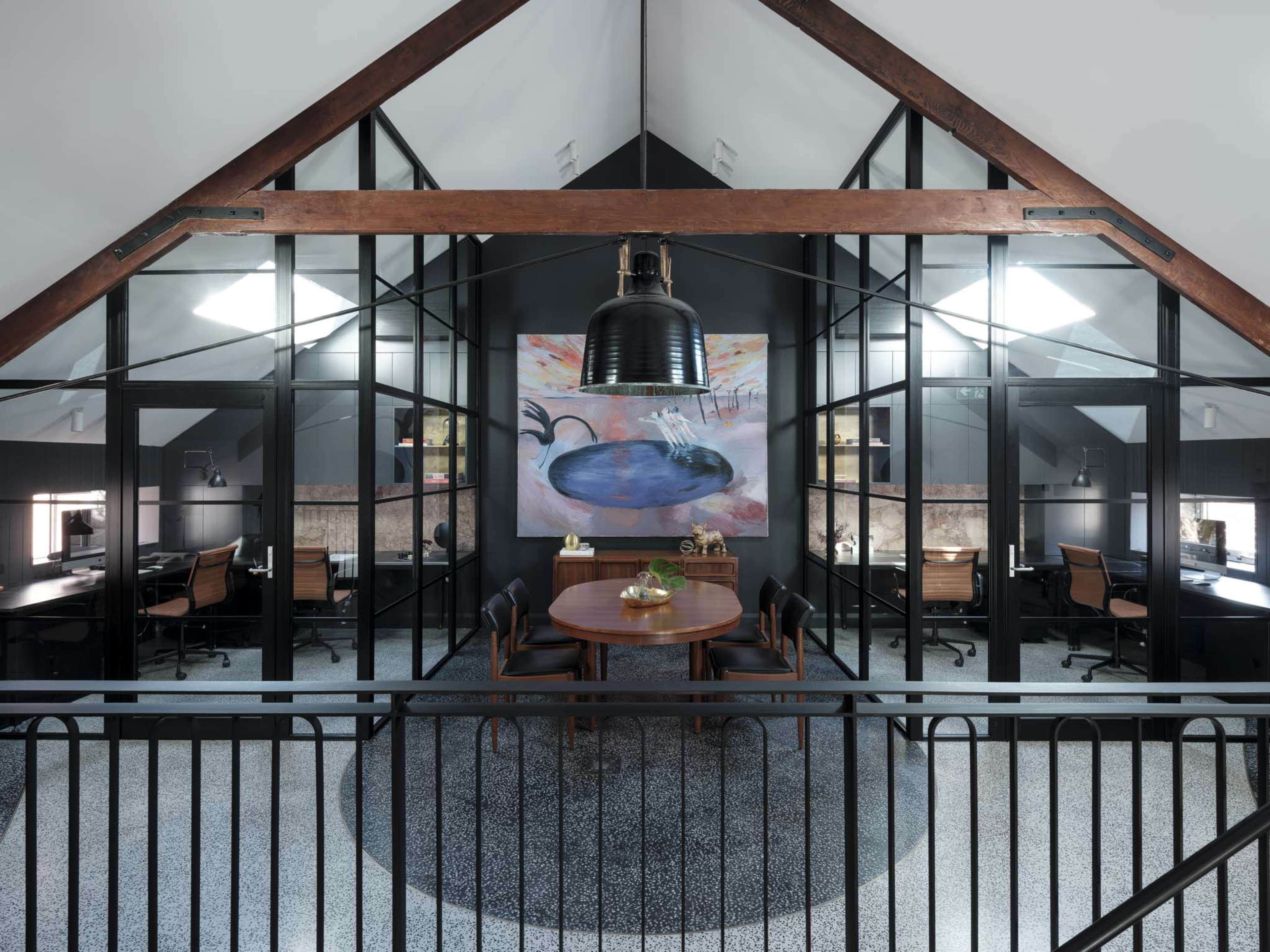 The image shows a modern office space in an attic with a central round table, surrounded by glass-paneled workstations and a large artwork on the wall.