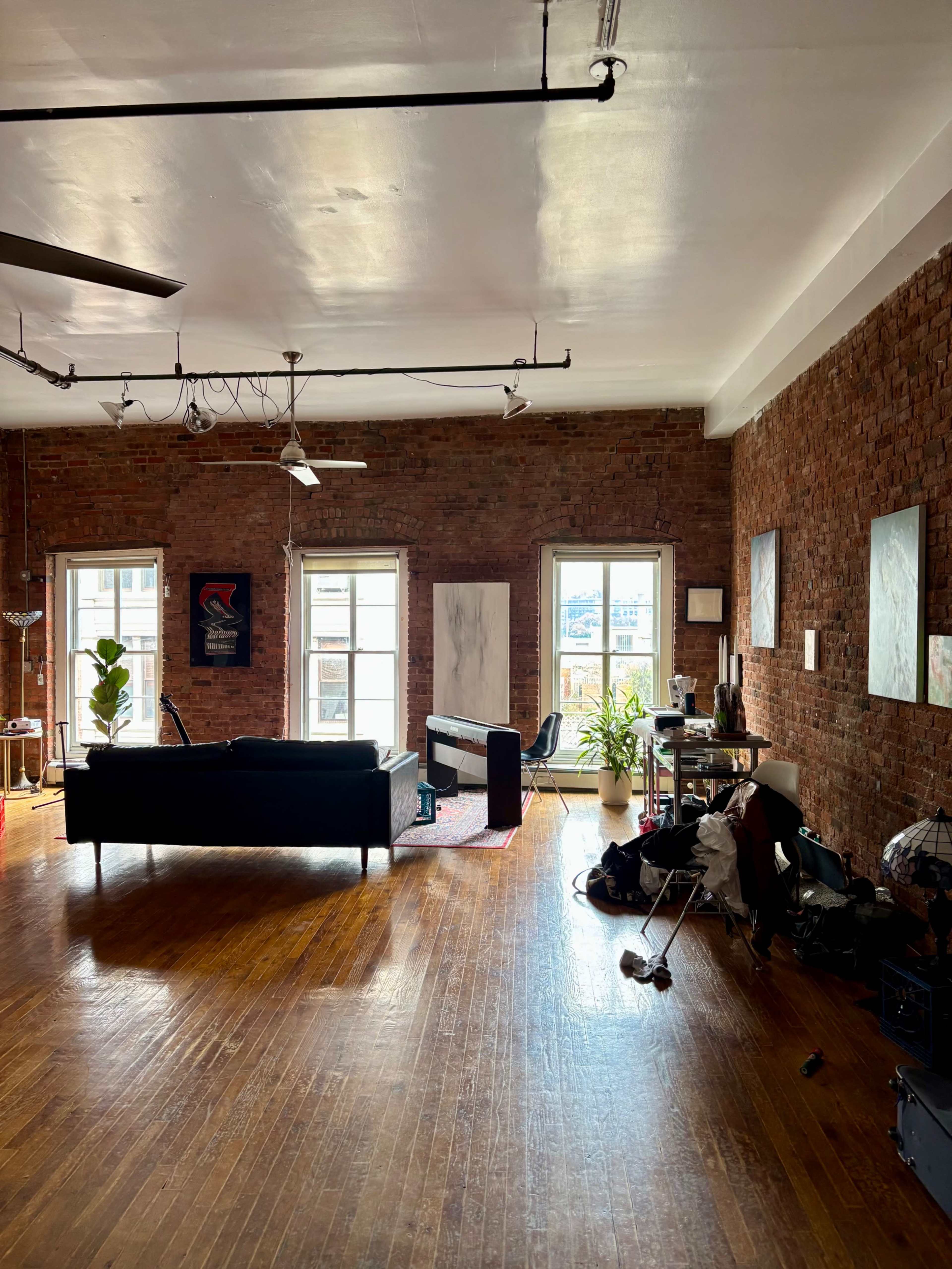 The image depicts a spacious living area with exposed brick walls, large windows, and a black sofa, along with various plants and artwork.