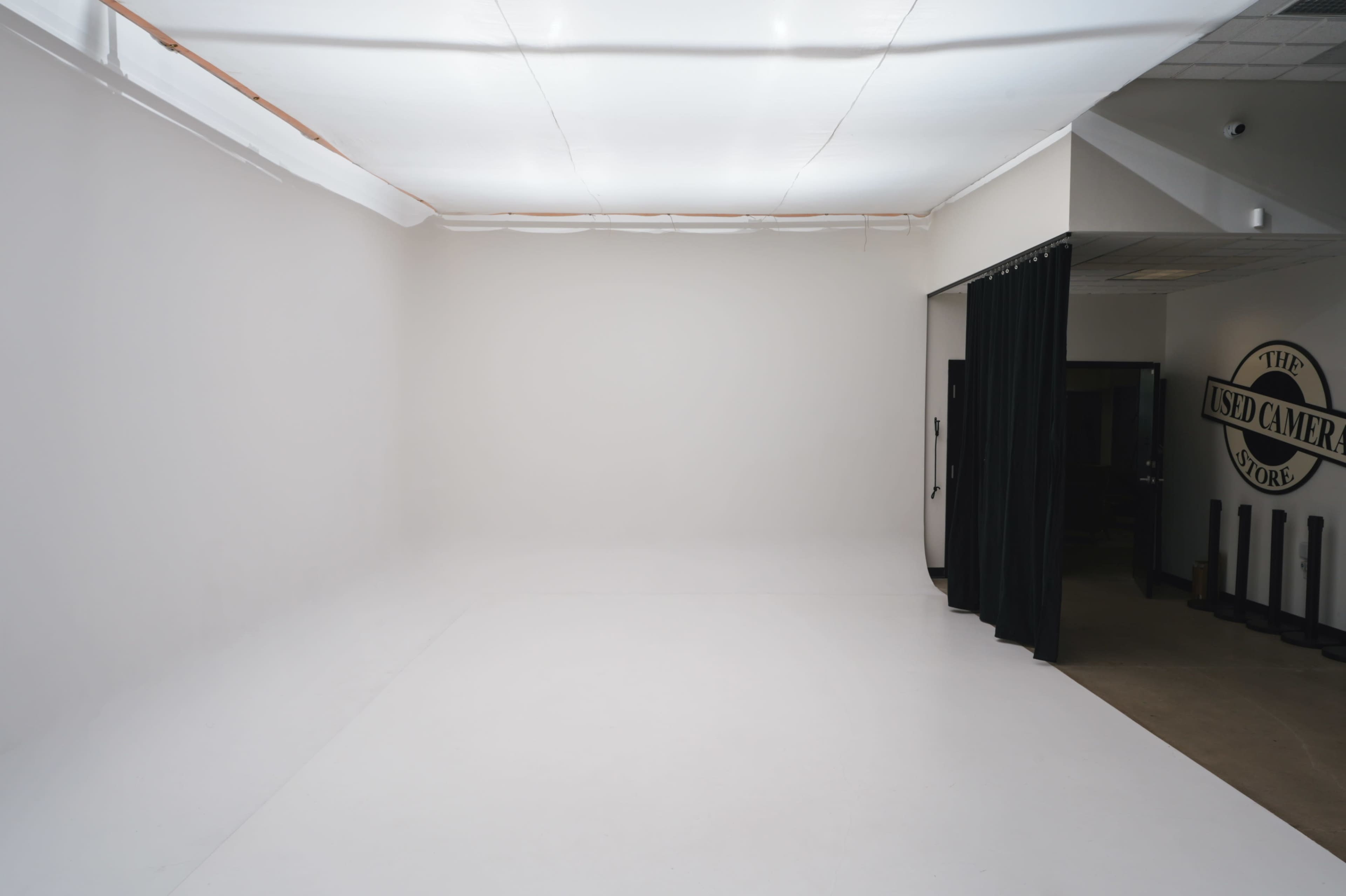 The image shows a well-lit, empty studio space with white walls and a black curtain on one side, leading to a doorway marked with "The Used Camera Store."