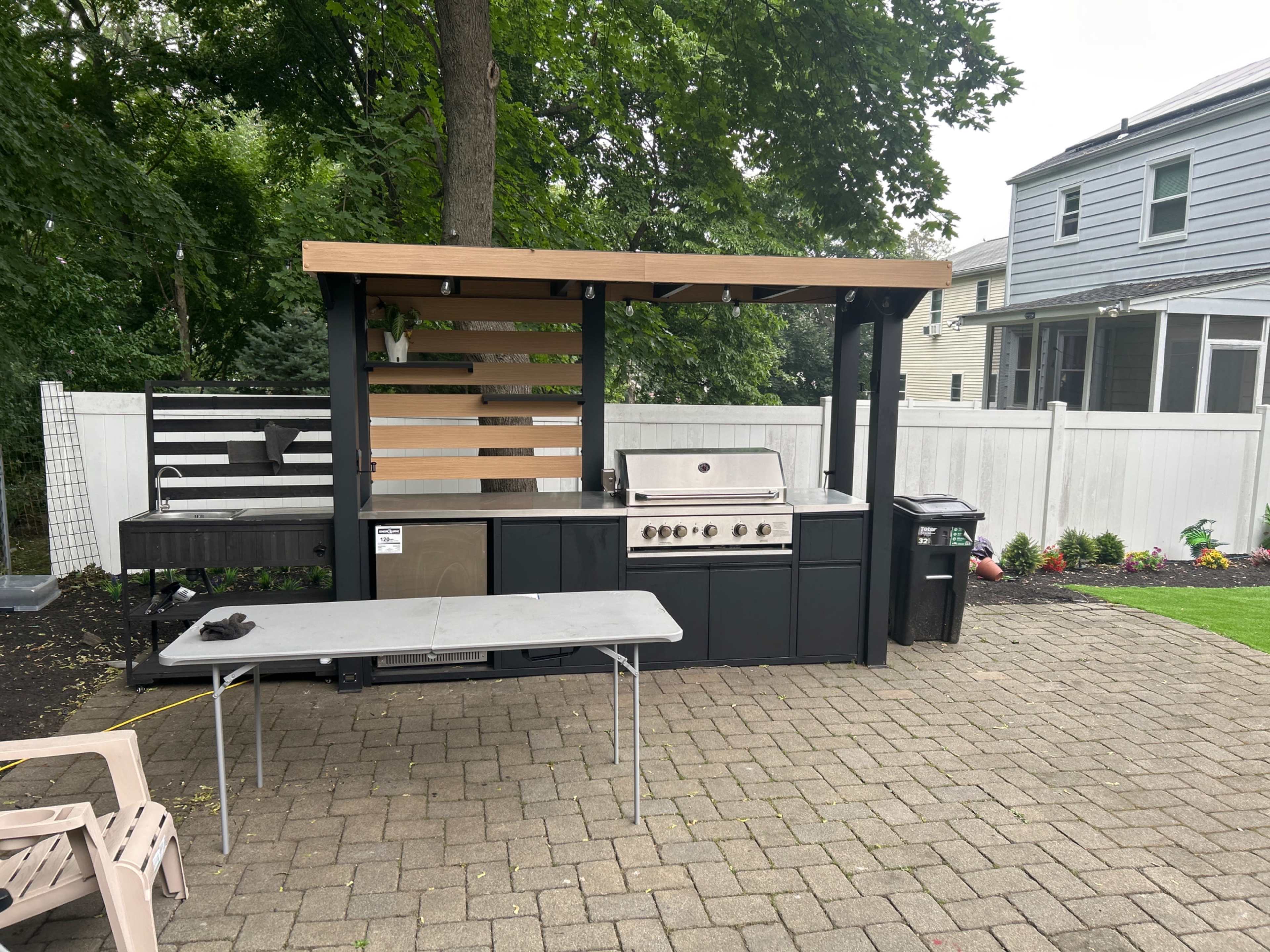 The image shows an outdoor kitchen setup featuring a grill, prep area, and a table, surrounded by a paved patio and residential buildings in the background.