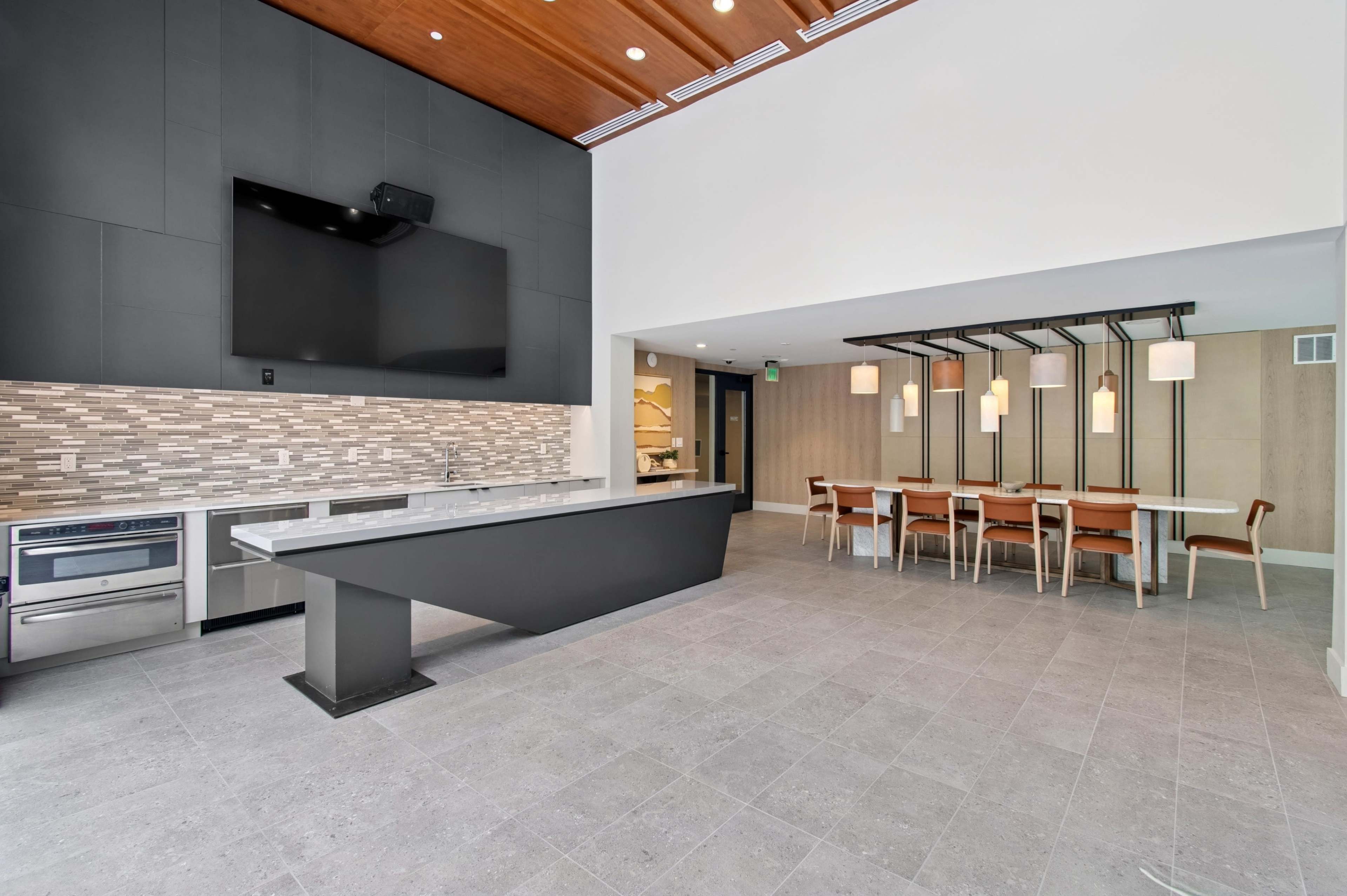 The image shows a modern kitchen and dining area featuring a large countertop, sleek cabinetry, a wall-mounted television, and a dining table with several chairs.