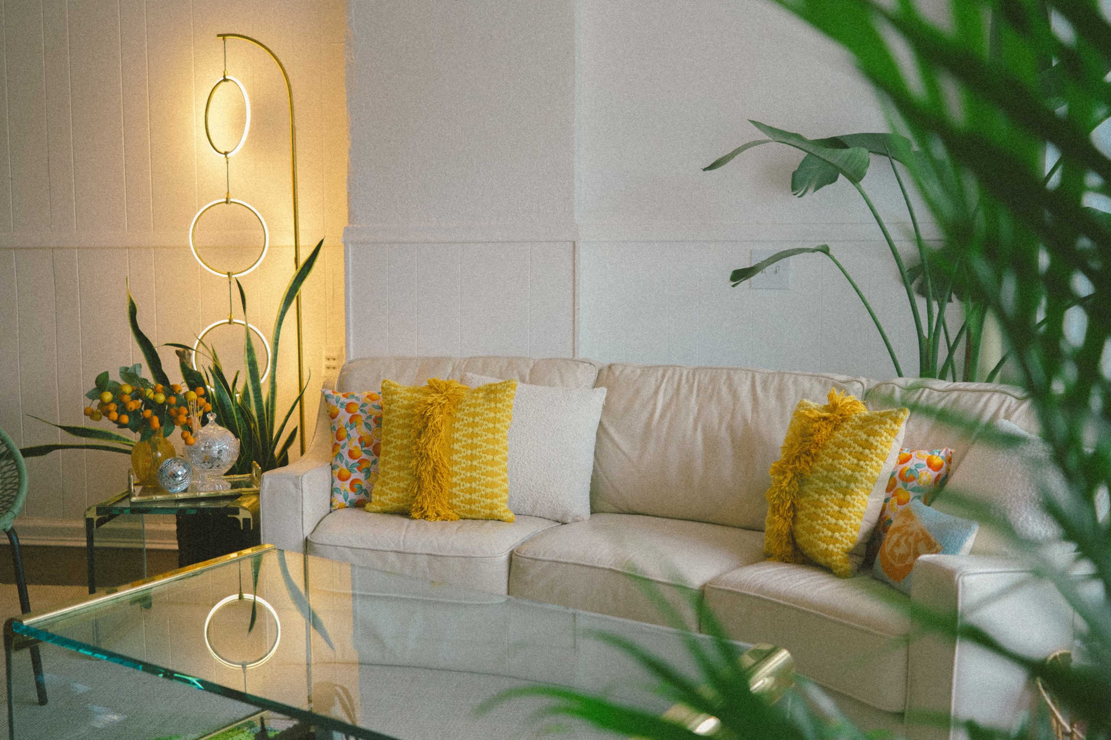 A modern living room features a light-colored sofa with decorative pillows, a glass coffee table, and potted plants, illuminated by a stylish floor lamp.