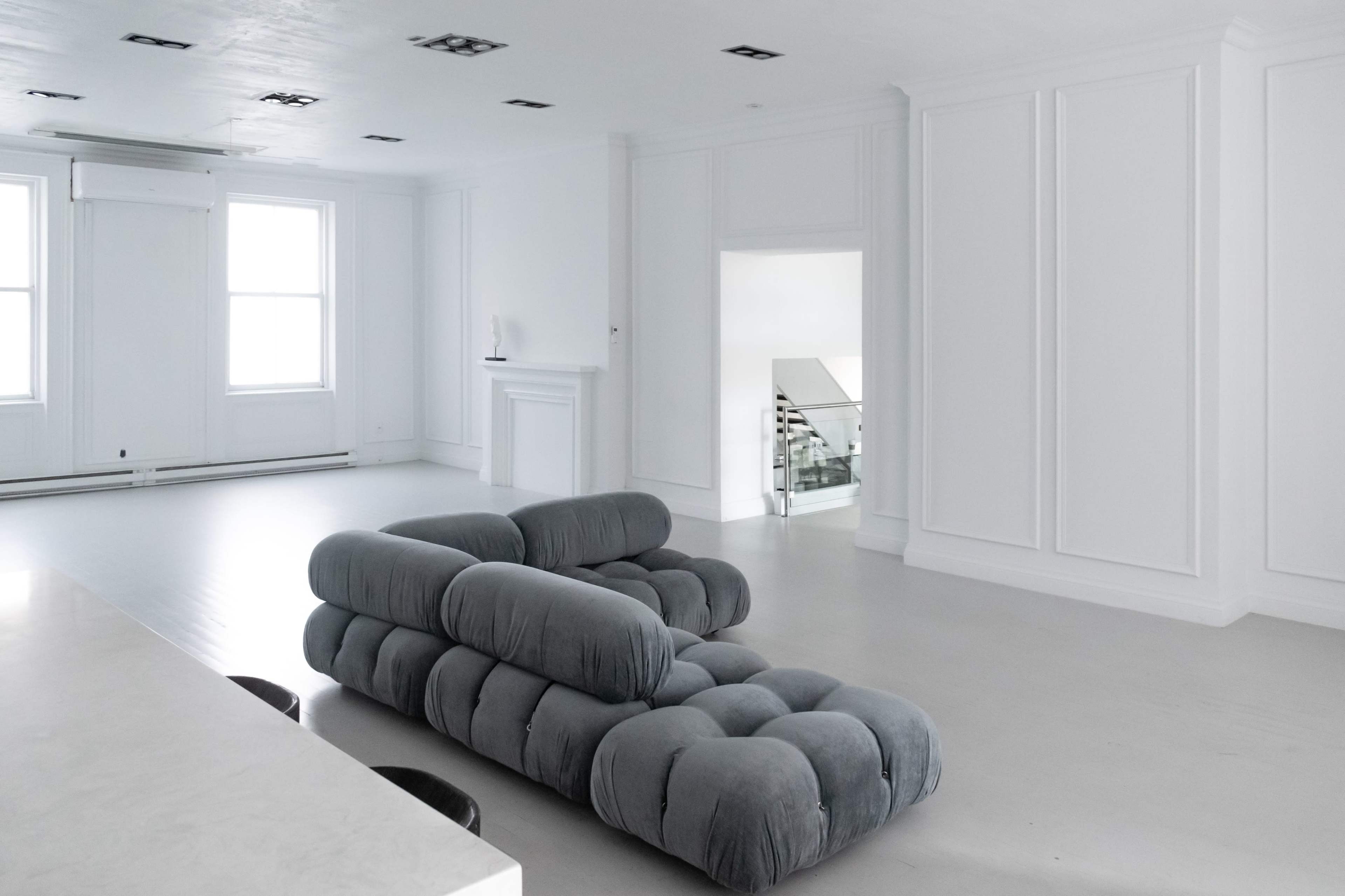 A spacious room with white walls and a gray tufted sofa positioned in the center, leading to a staircase at the far end.