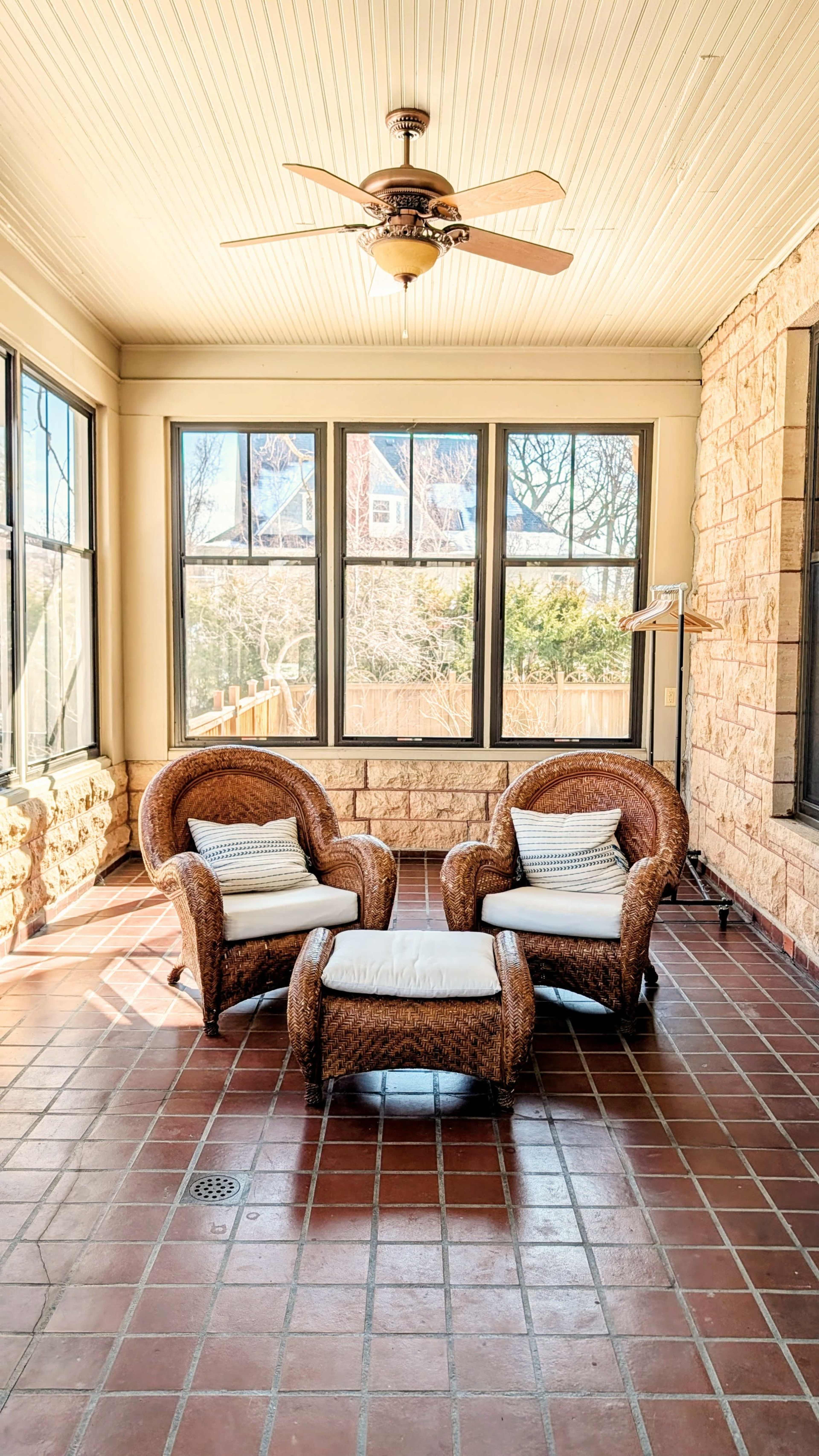The image shows a sunlit sunroom featuring two wicker chairs with cushions and a small table between them, surrounded by large windows.