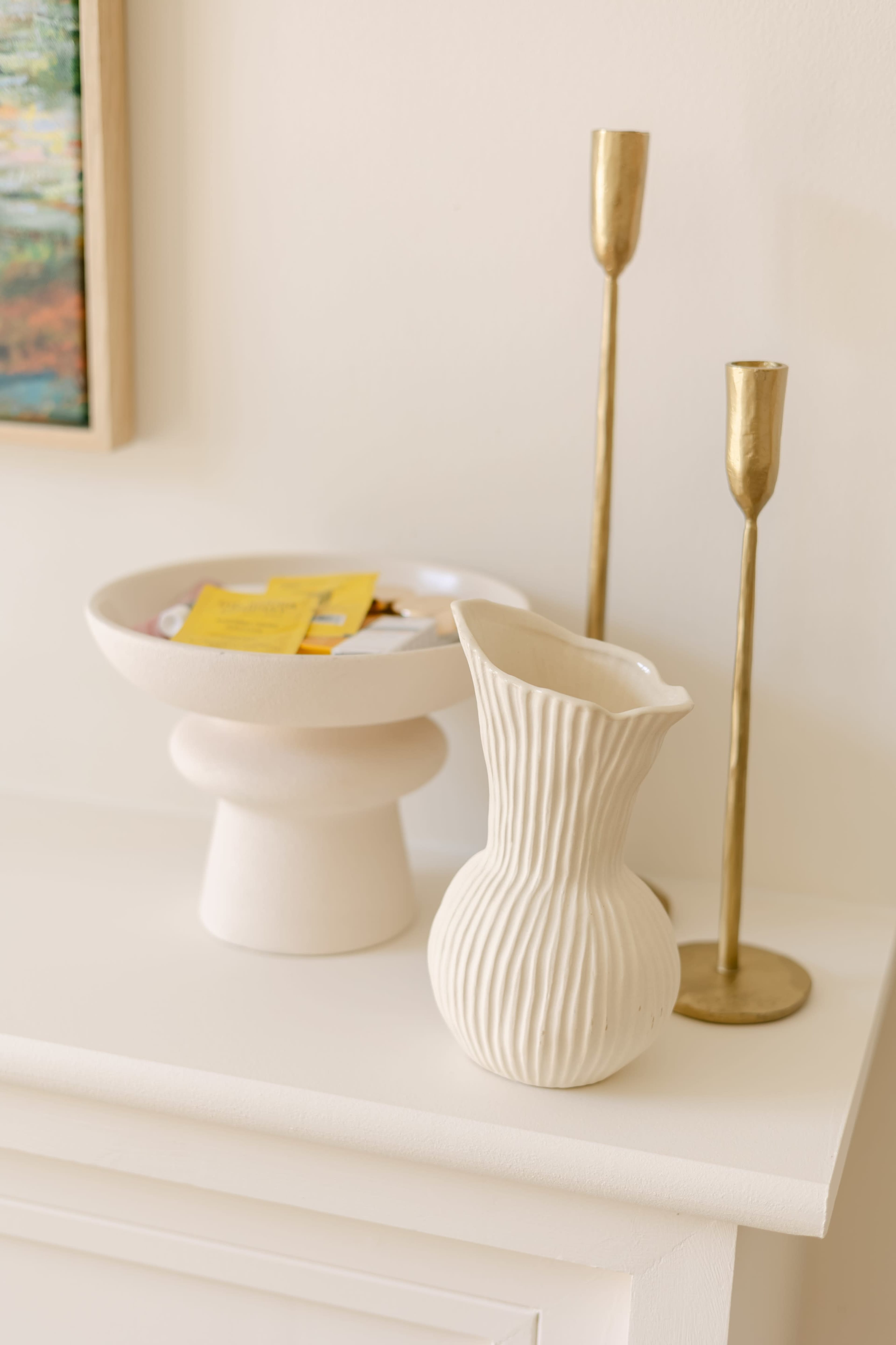 The image features a minimalist arrangement on a white surface, including a textured white vase, a round bowl filled with items, and two tall gold candle holders.