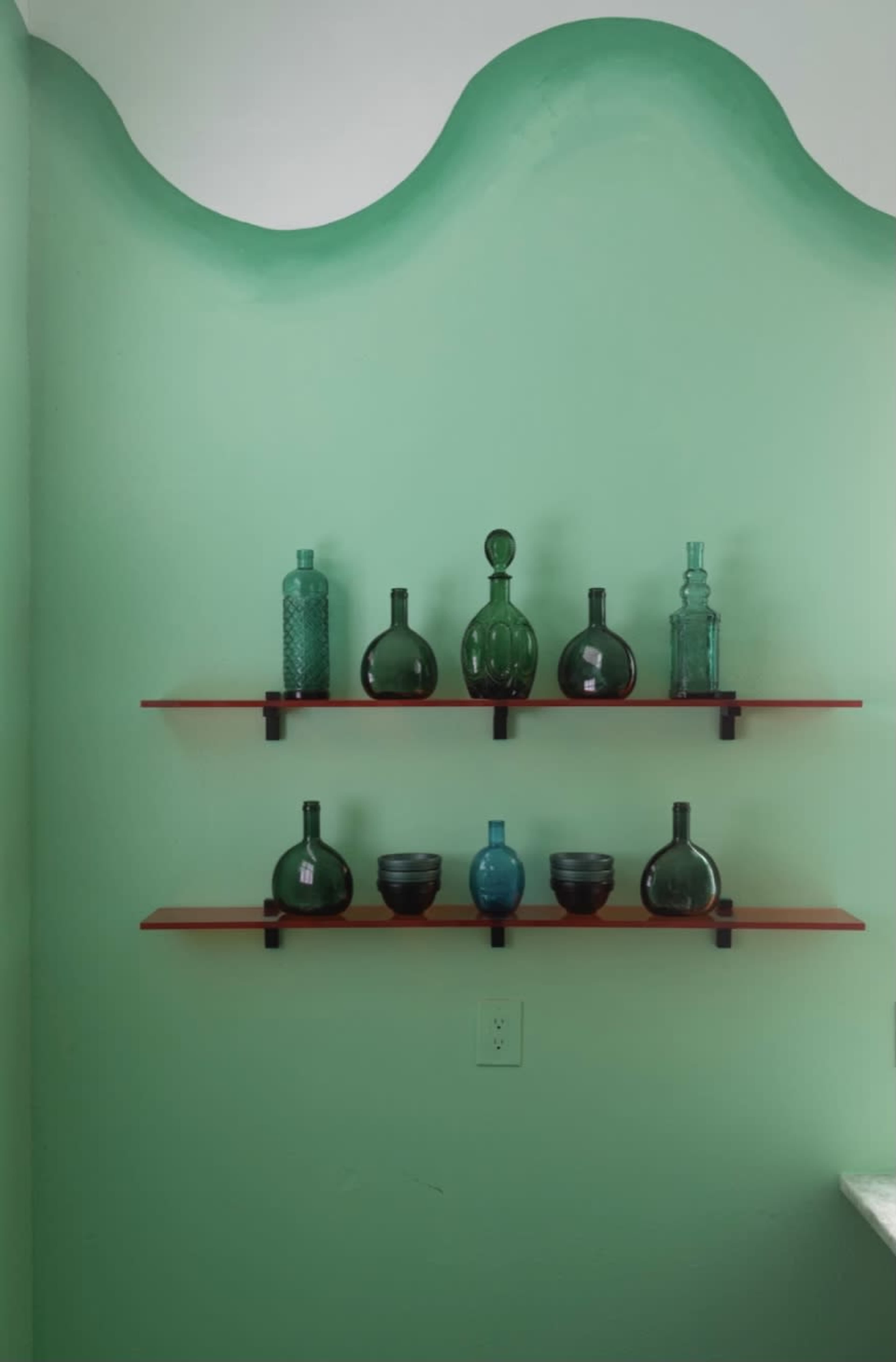 Three shelves hold various glass bottles and bowls against a light green wall with a wavy top edge.