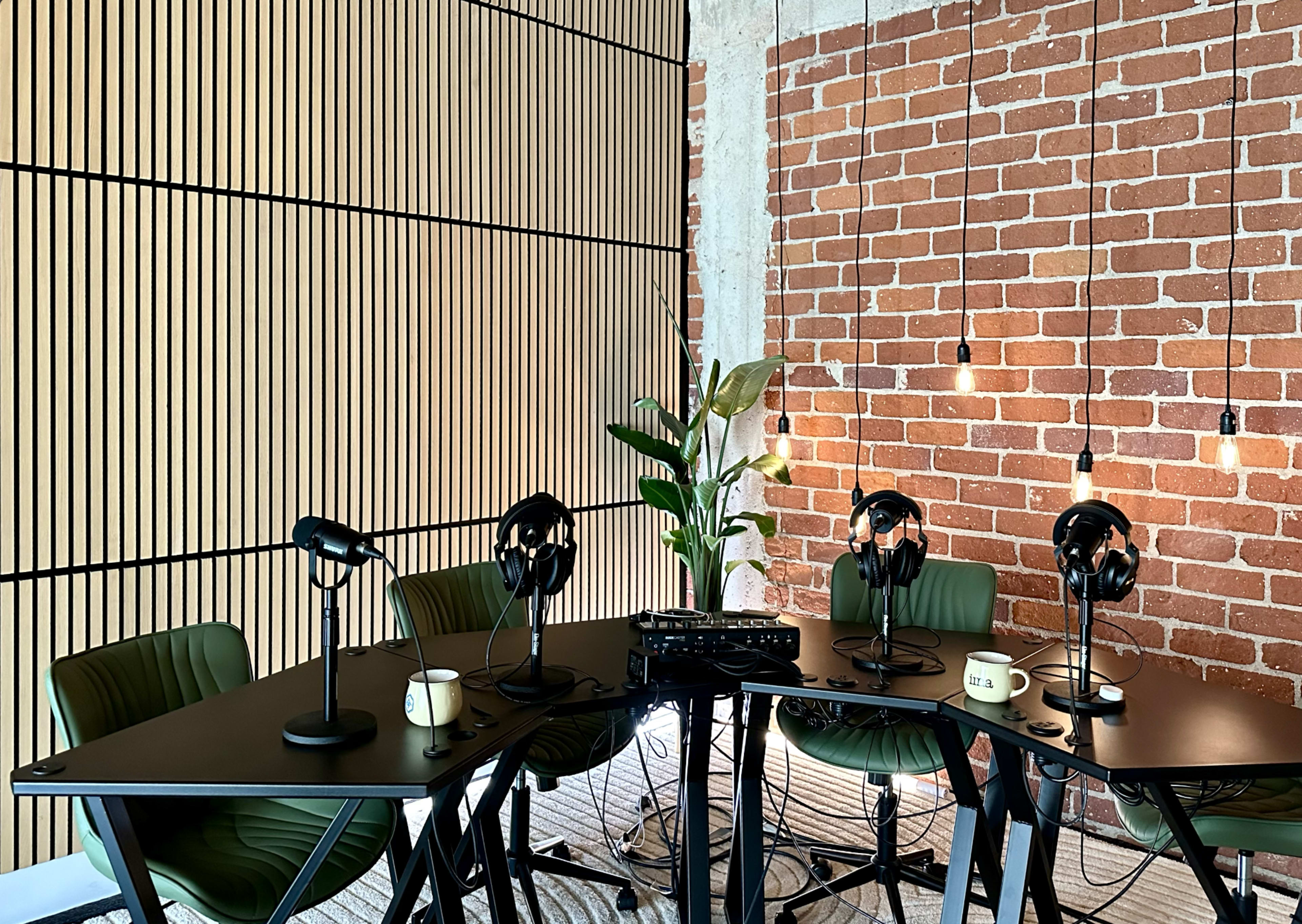 A podcast studio features a table with four green chairs, microphones, and hanging light fixtures, set against a brick wall and wooden paneling.