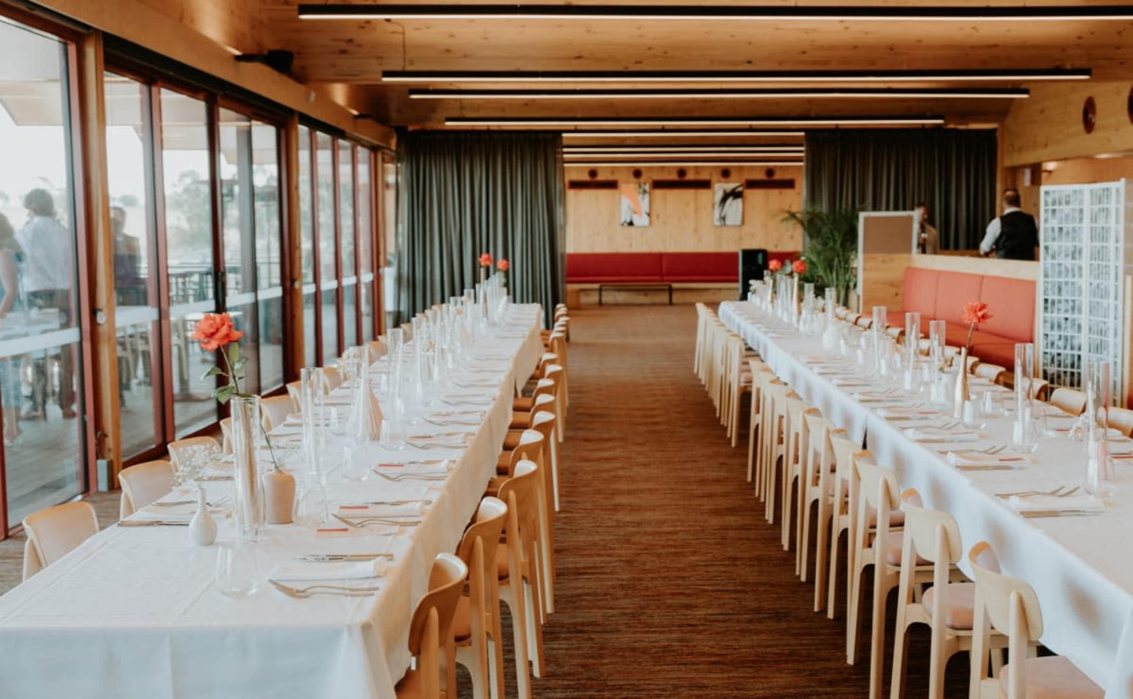 The image shows a spacious dining area with two long tables set with white tablecloths, each featuring a single flower in a vase and neatly arranged cutlery.