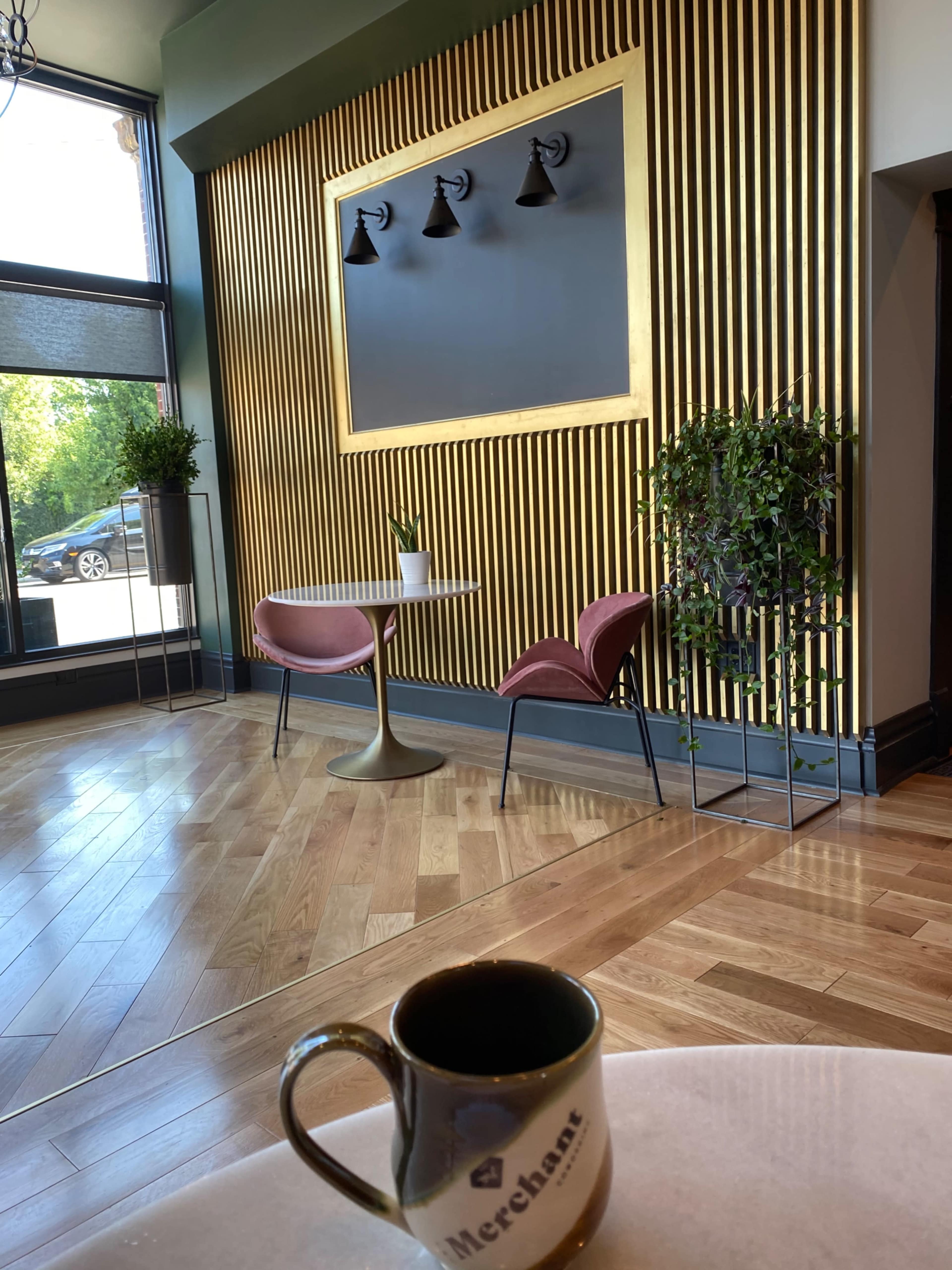 The image shows a cozy café interior with wooden flooring, a decorative wall featuring vertical gold slats and four light fixtures, along with two pink chairs and a small table, while a coffee cup sits on the table.