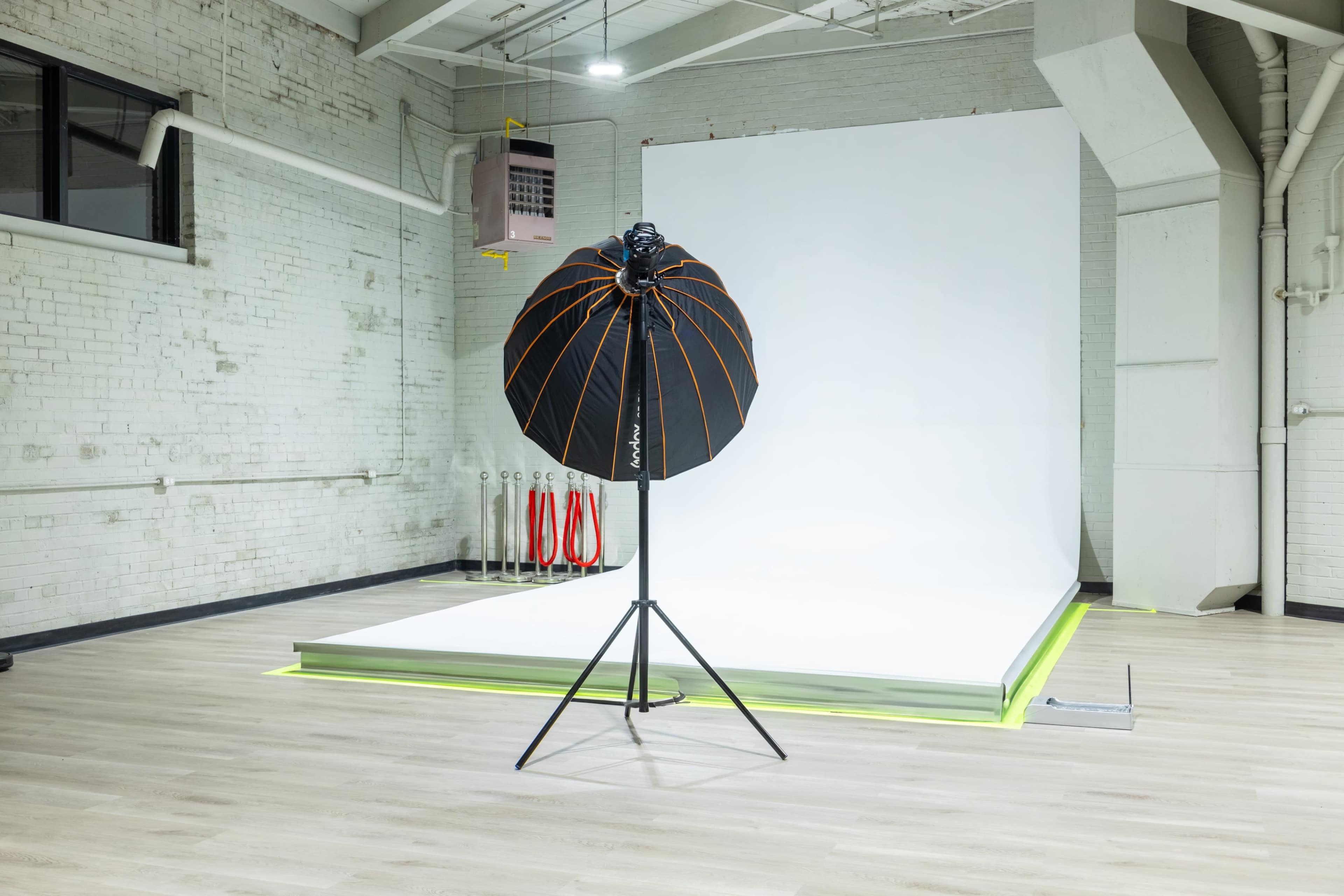 The image shows a photography studio with a white backdrop, a light stand equipped with a large umbrella, and a wooden floor.