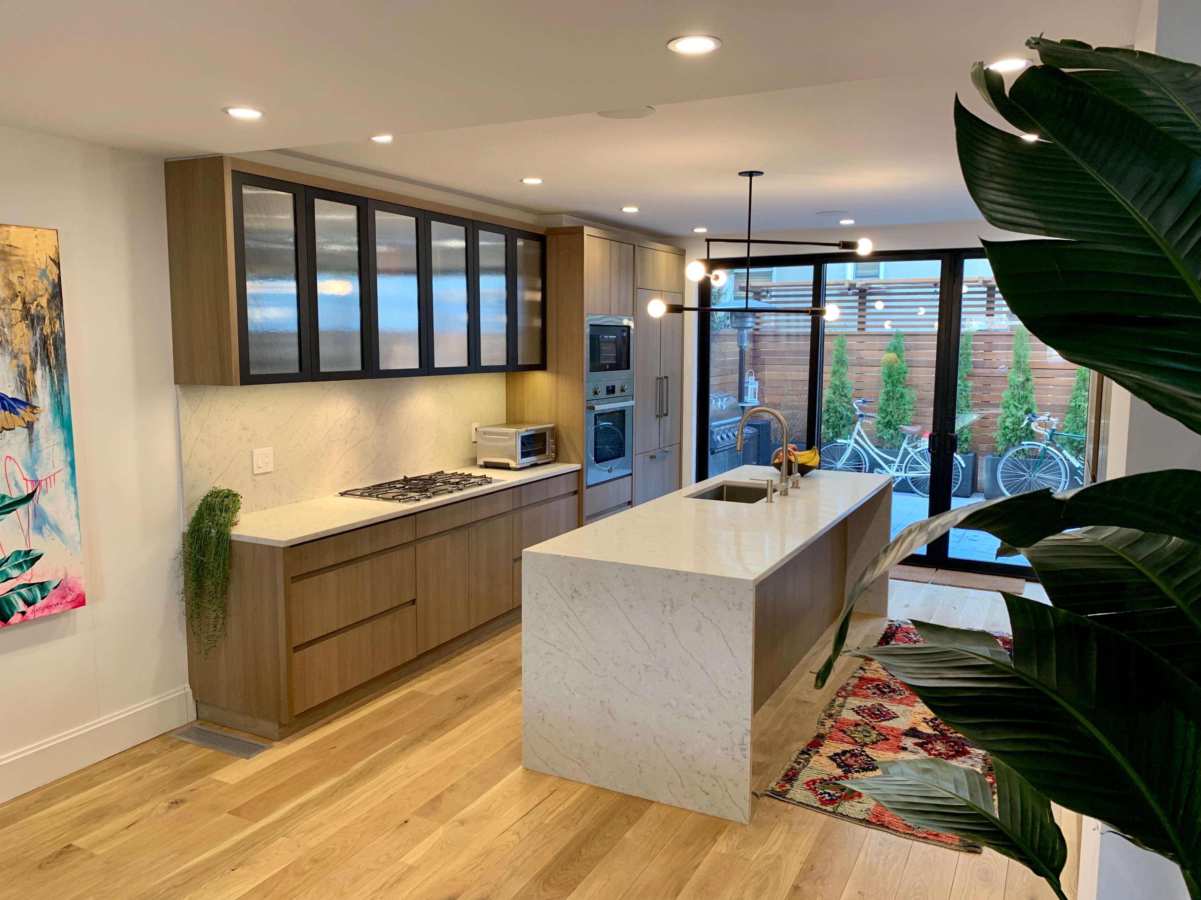 A modern kitchen features wooden cabinets, marble countertops, and large windows opening to an outdoor space with bicycles visible.