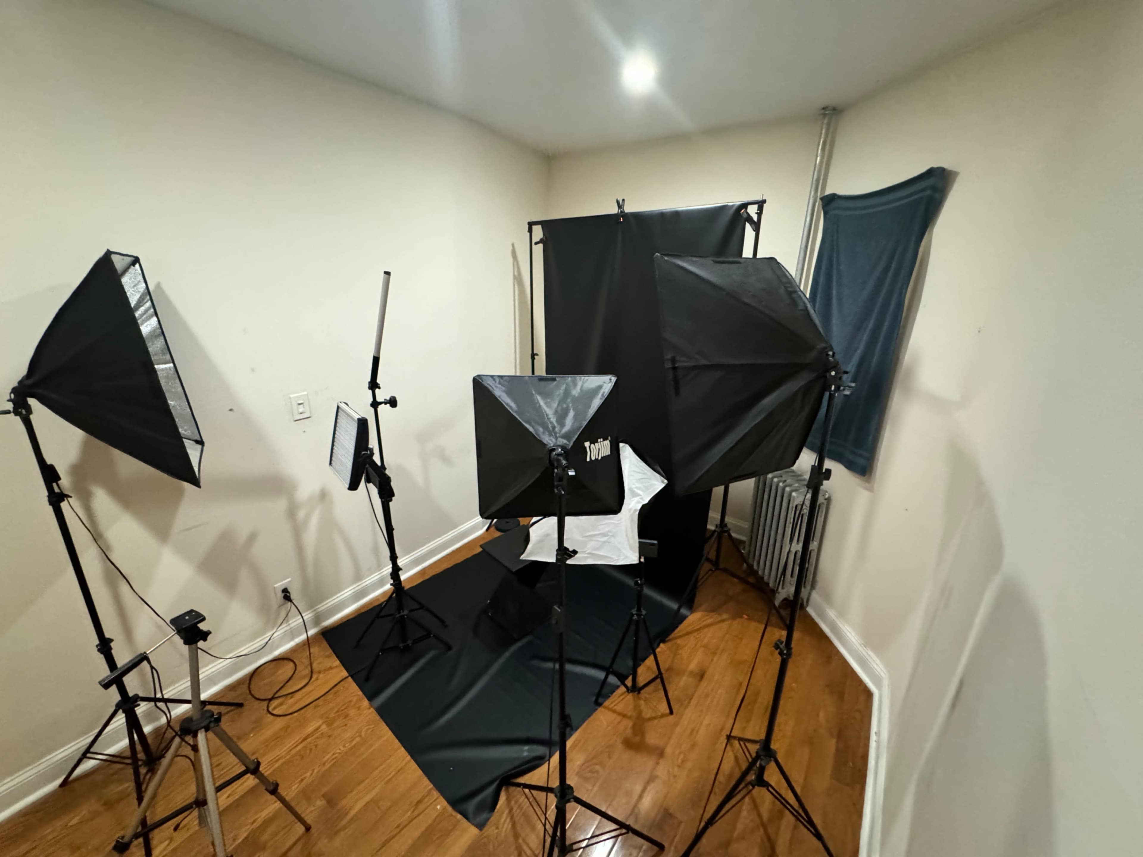 The image shows a small photography studio setup with two softbox lights, a backdrop, and a tripod in the corner of a room.