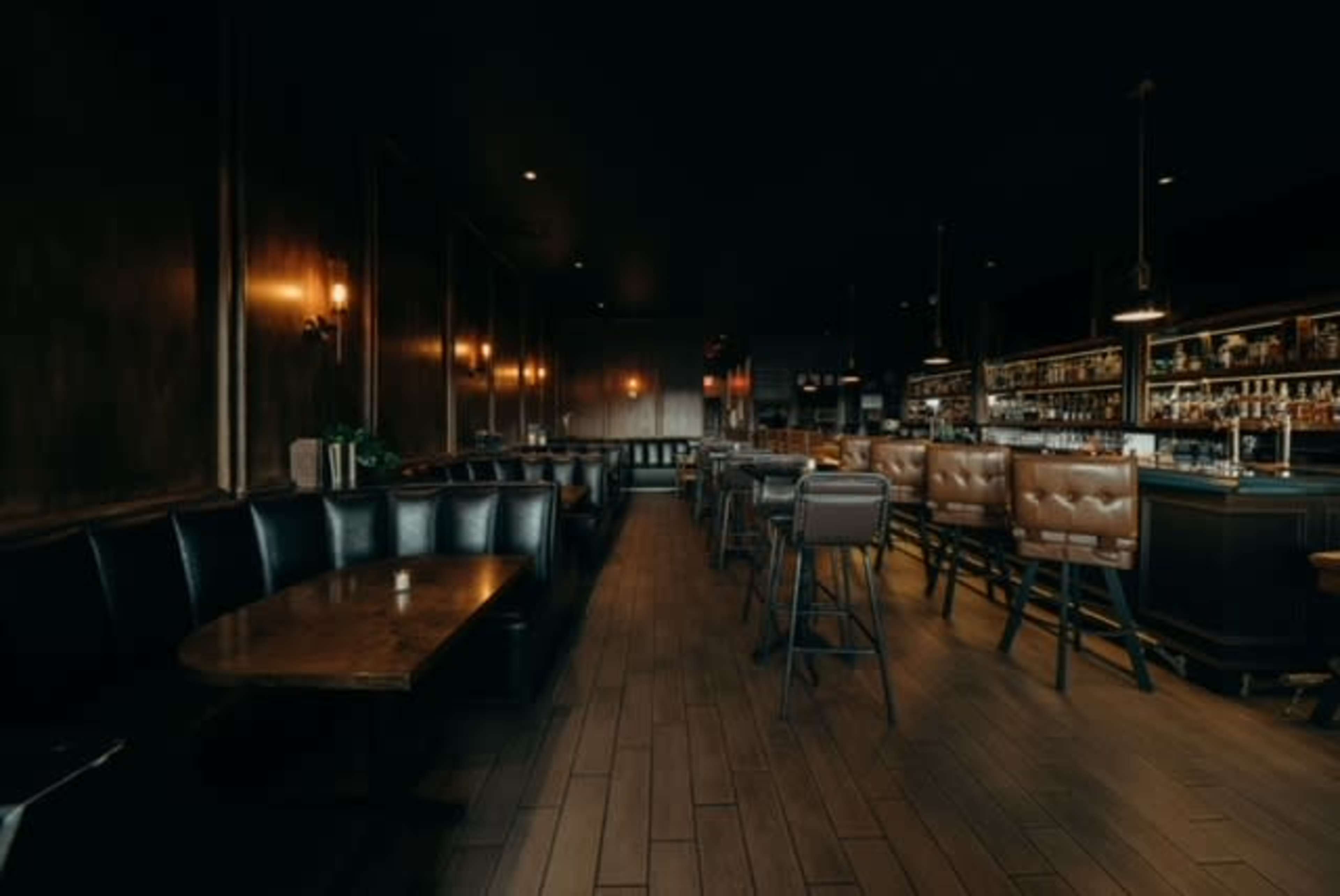 The interior of a dimly lit bar features leather seating, wooden tables, and a long bar with shelves of liquor in the background.