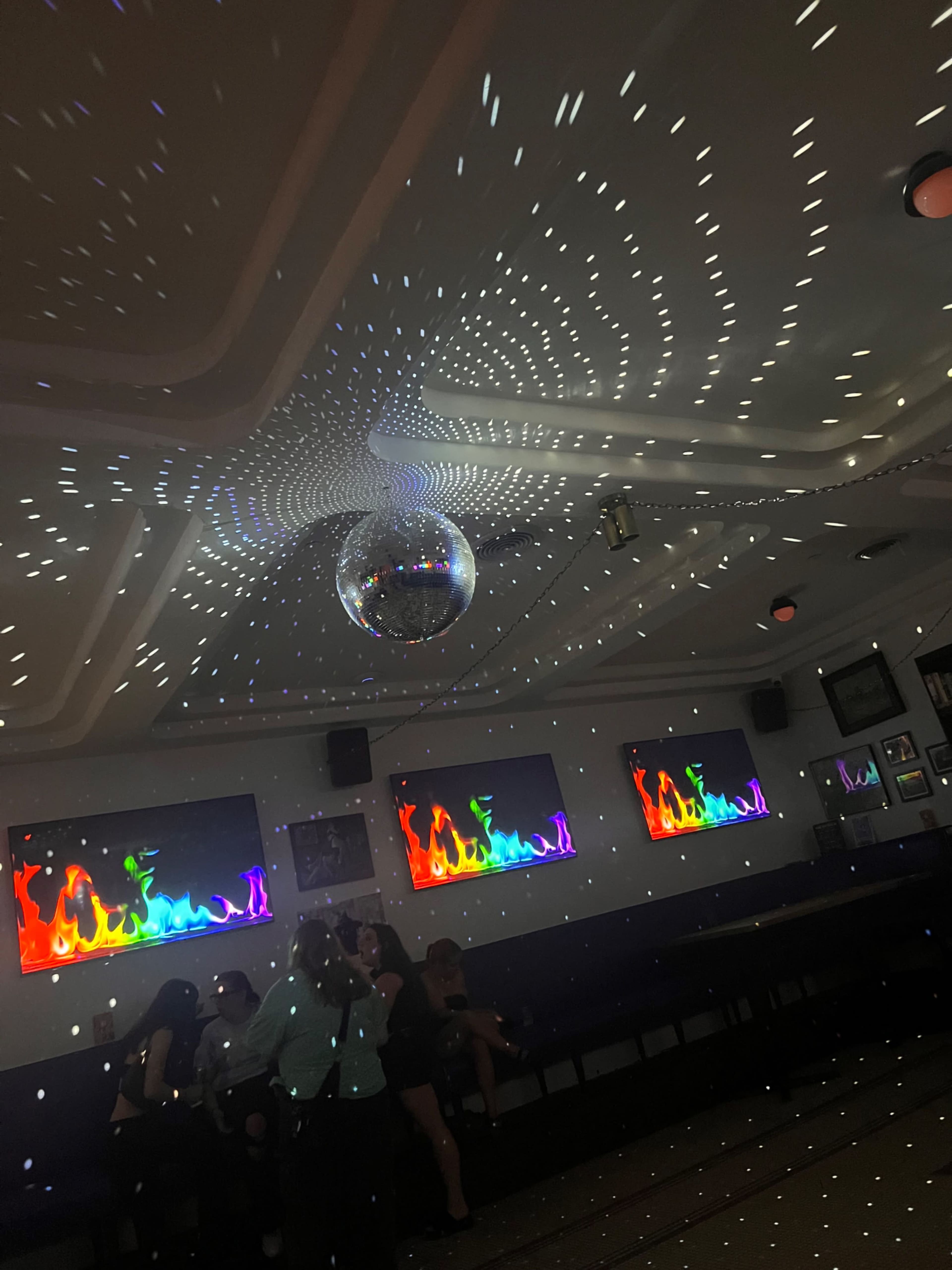 A disco ball reflects multicolored lights around a room with digital screens displaying vibrant graphics and a group of people sitting on benches.