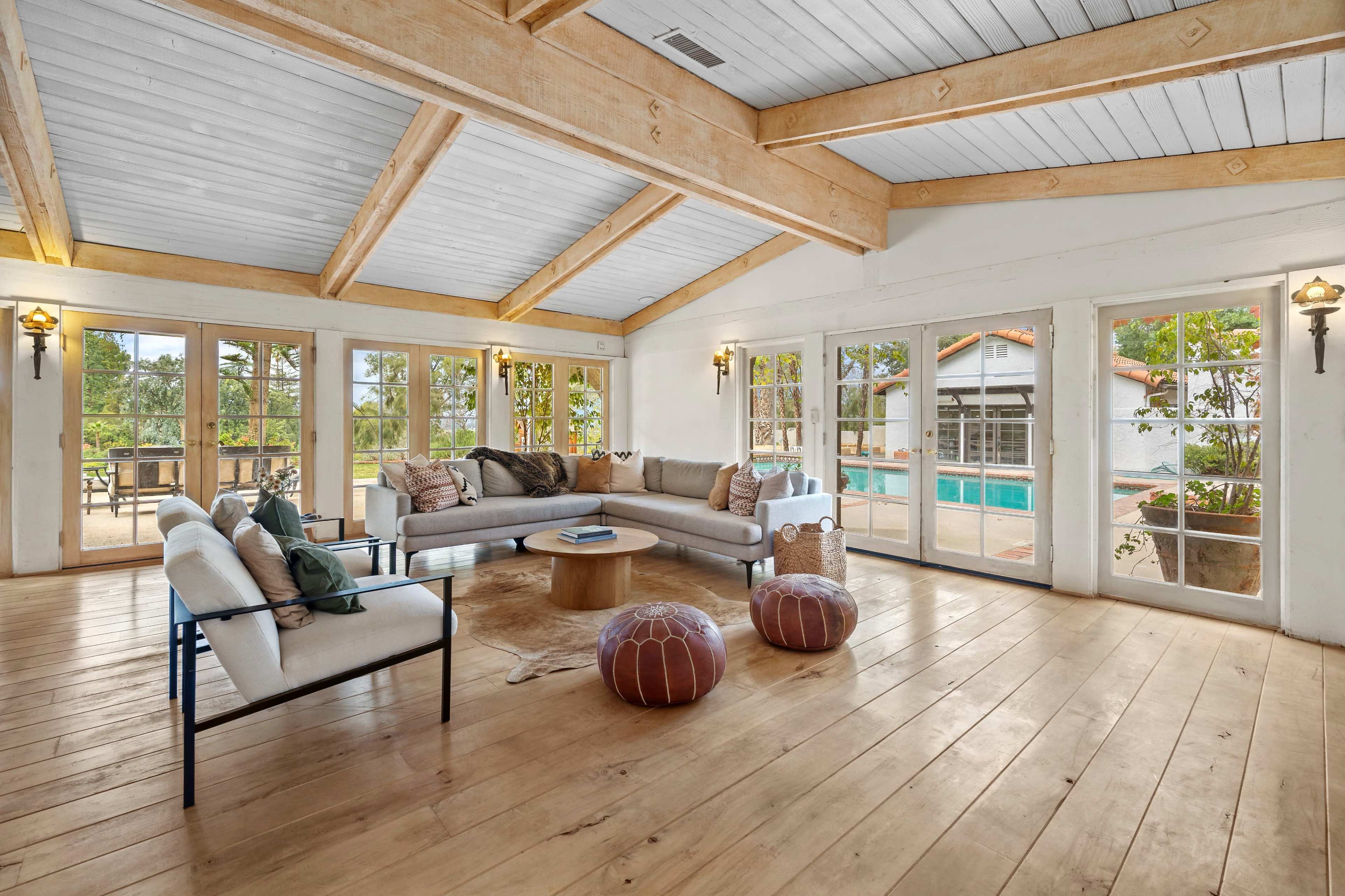 A spacious living room features large windows, wooden beams, and a view of a pool area outside.