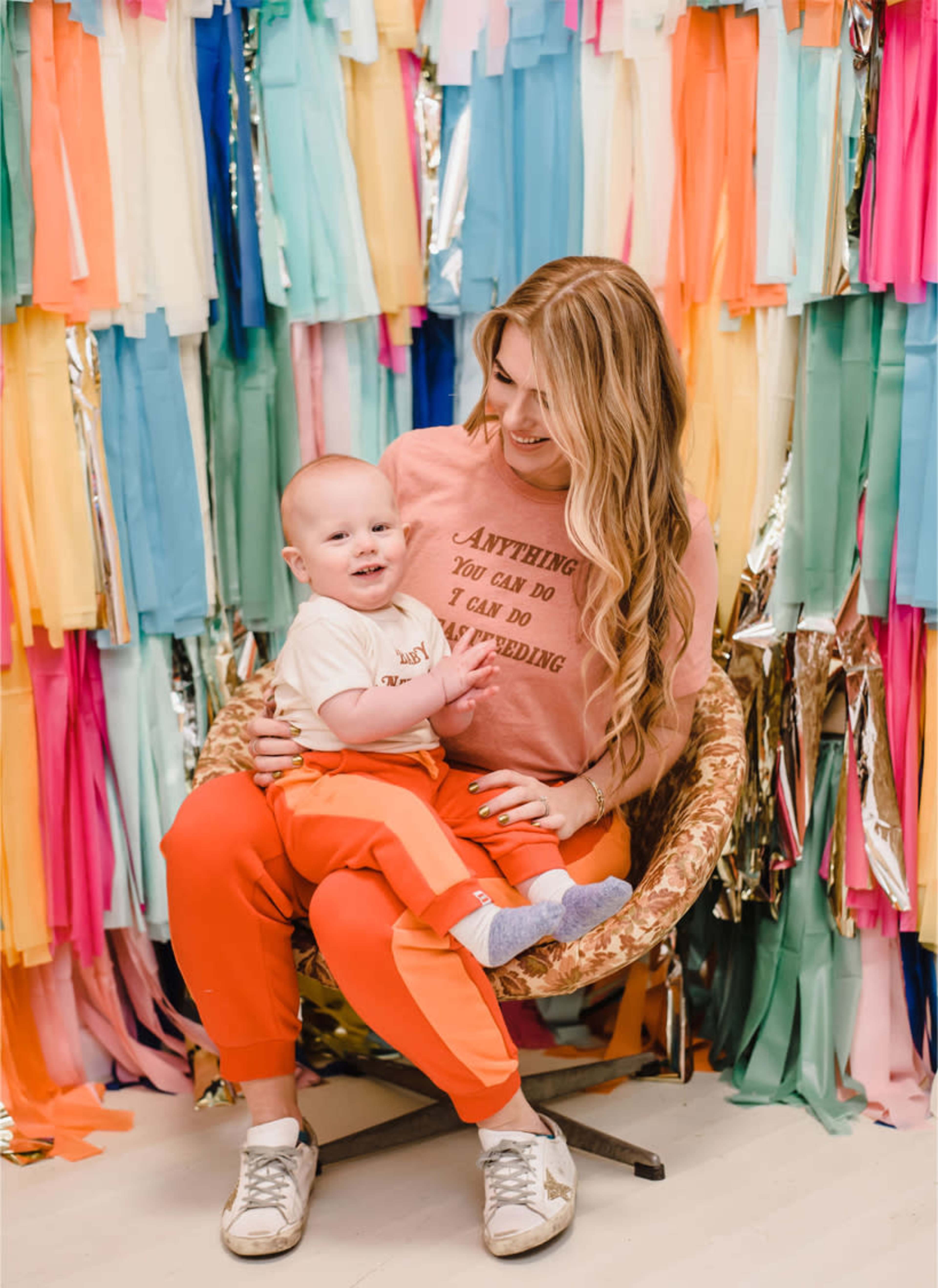 A woman sits in a chair with a baby on her lap, surrounded by a colorful backdrop of fabric strips.