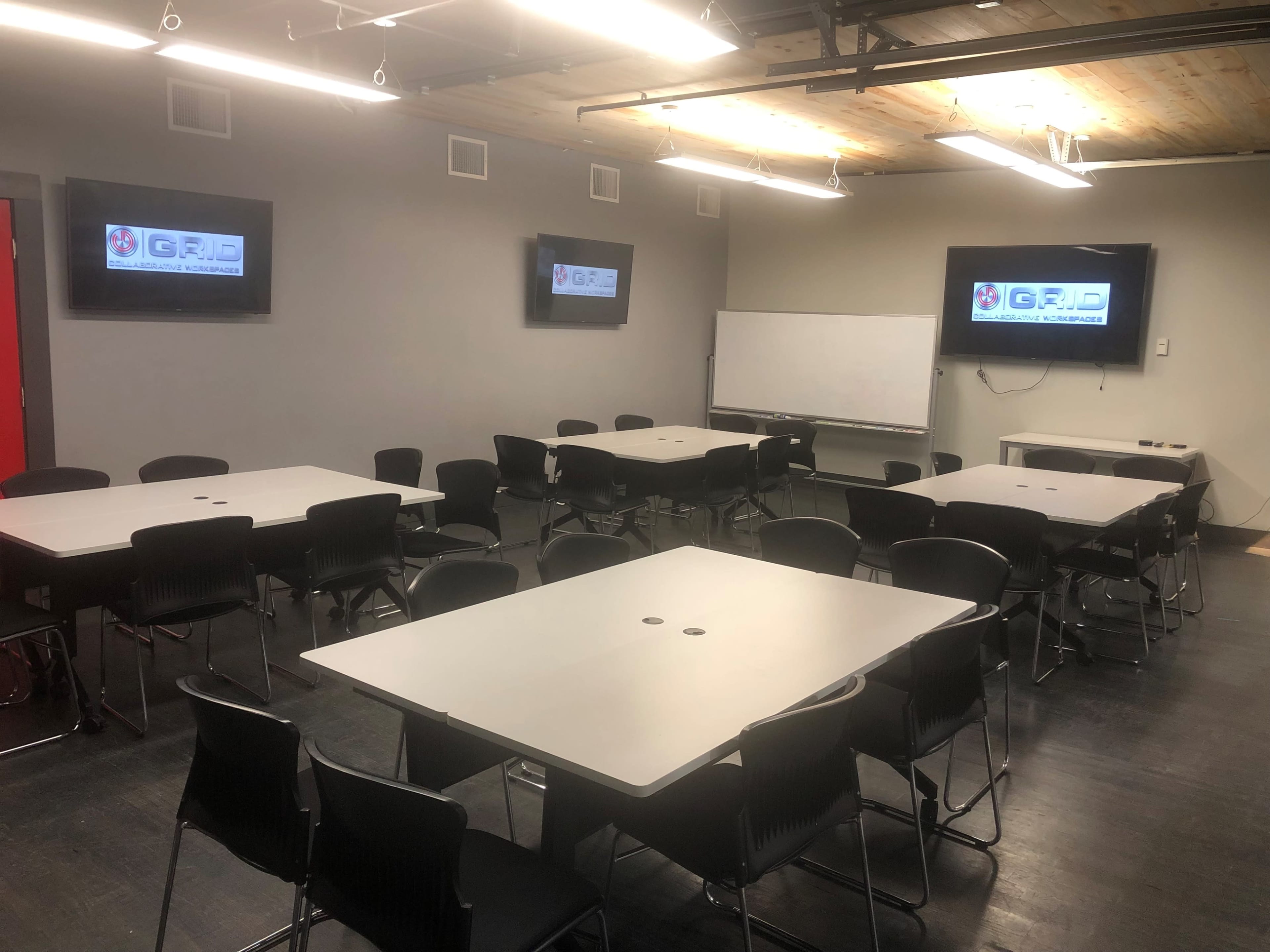 The image shows a meeting room with several tables arranged and chairs placed around them, accompanied by two monitors on the walls and a whiteboard.