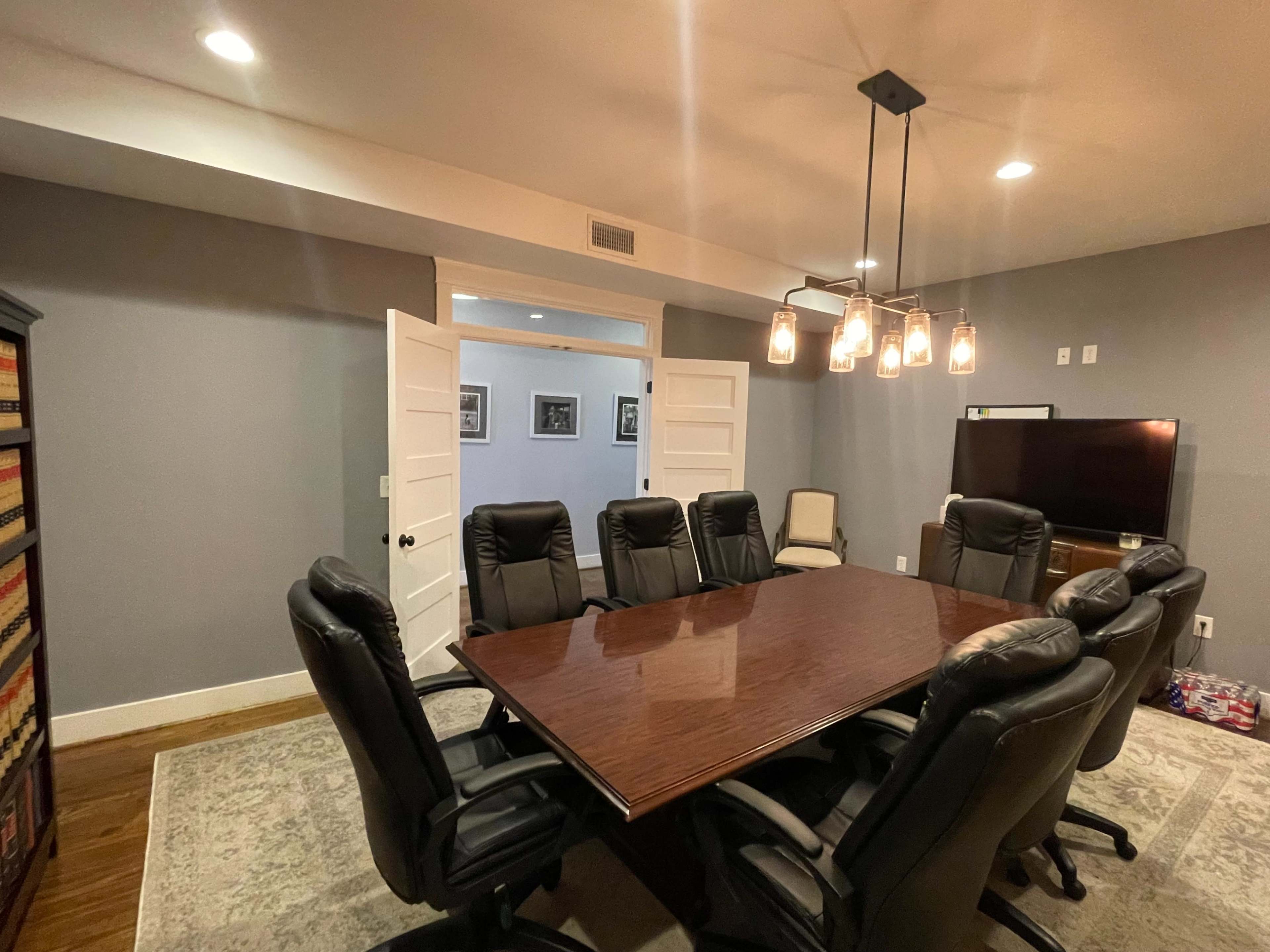 A conference room features a large wooden table surrounded by six black ergonomic chairs, with two doors leading to another area and a television mounted on the wall.