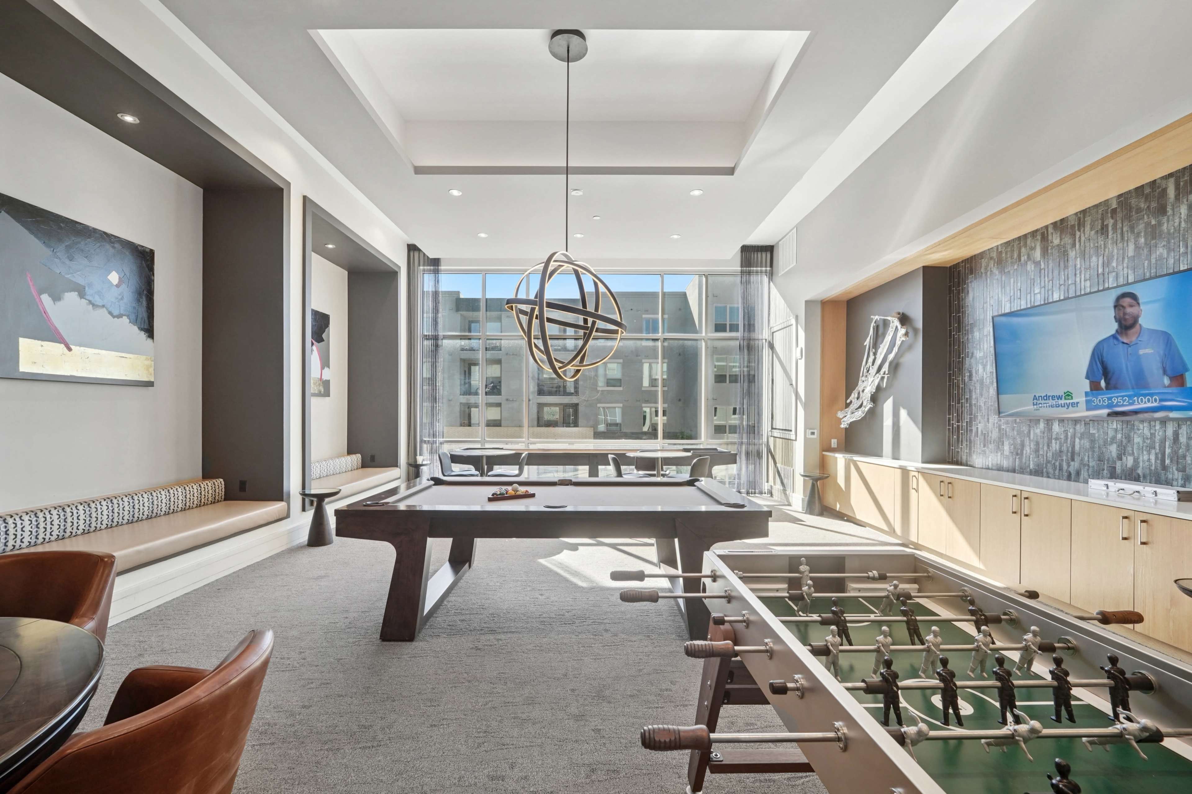 The image shows a modern recreational room featuring a billiard table, foosball table, large windows, and contemporary artwork on the walls.