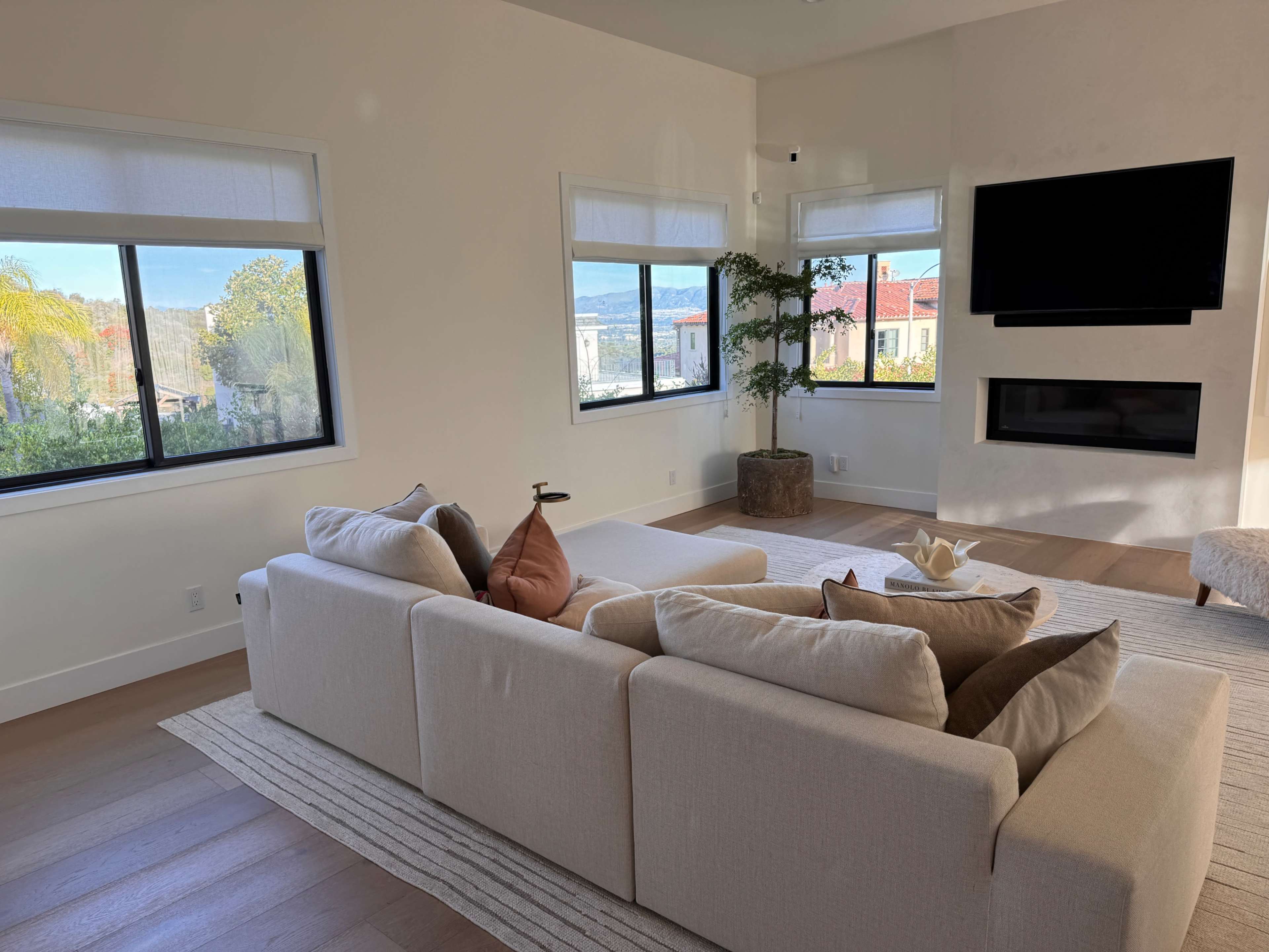 A modern living room features a light-colored sectional sofa, a large window with scenic views, and a mounted TV above a fireplace.